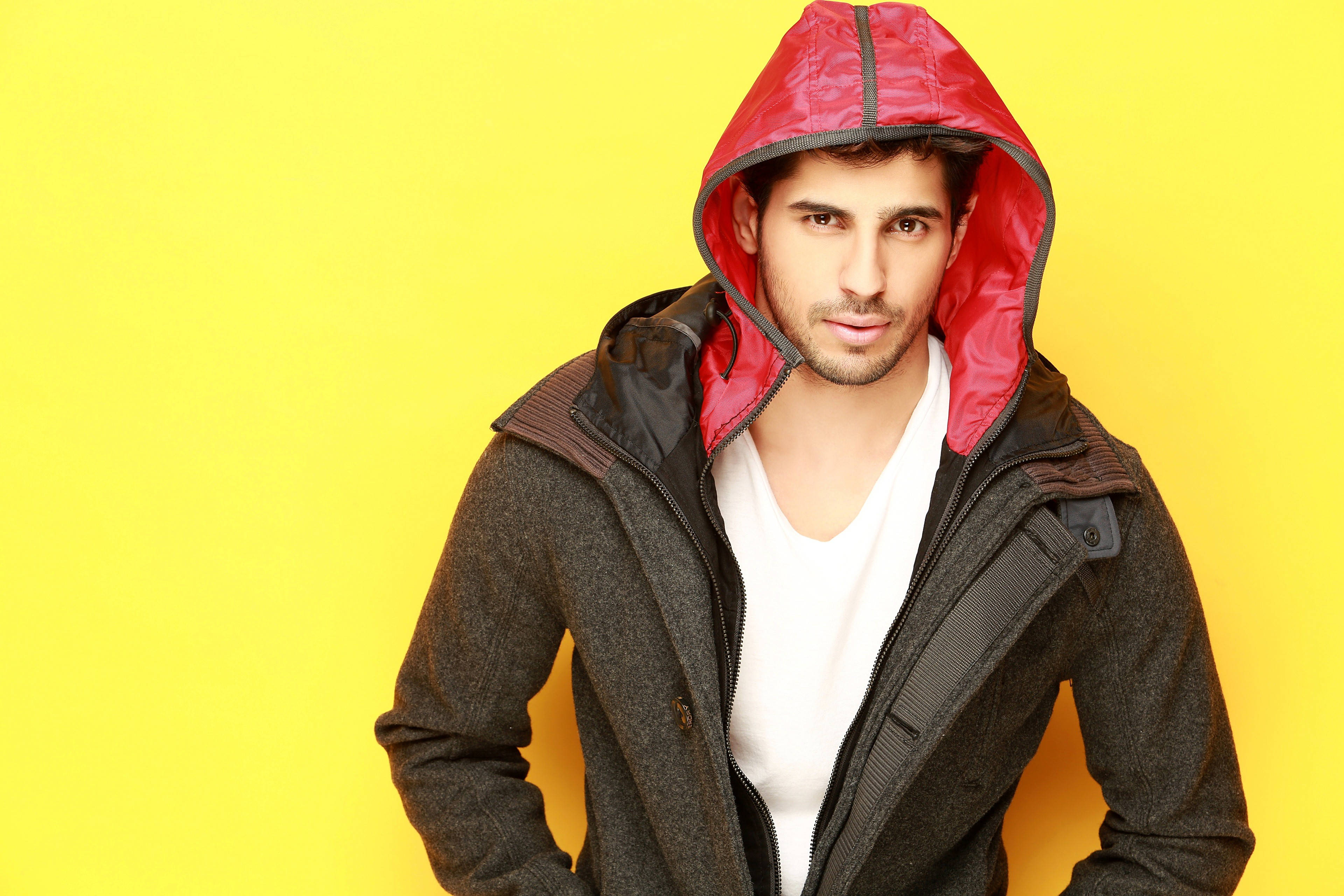 Sidharth Malhotra 桌布 壁紙 — 一個穿著紅色內裡連帽外套的男人在鮮黃色背景前自信地擺姿勢。