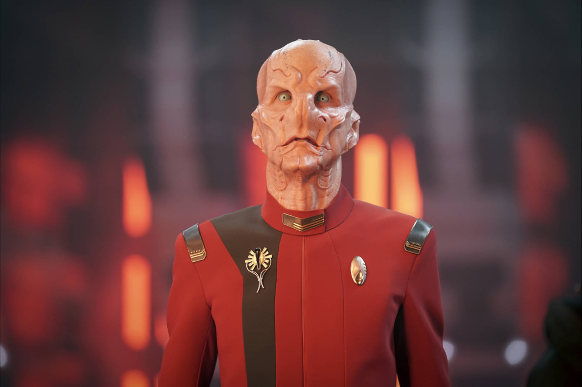 Discovery 桌布 壁紙 — 一個穿著紅色制服的人形外星人站在模糊的火焰背景前，伴隨著發光的橙色燈光。
