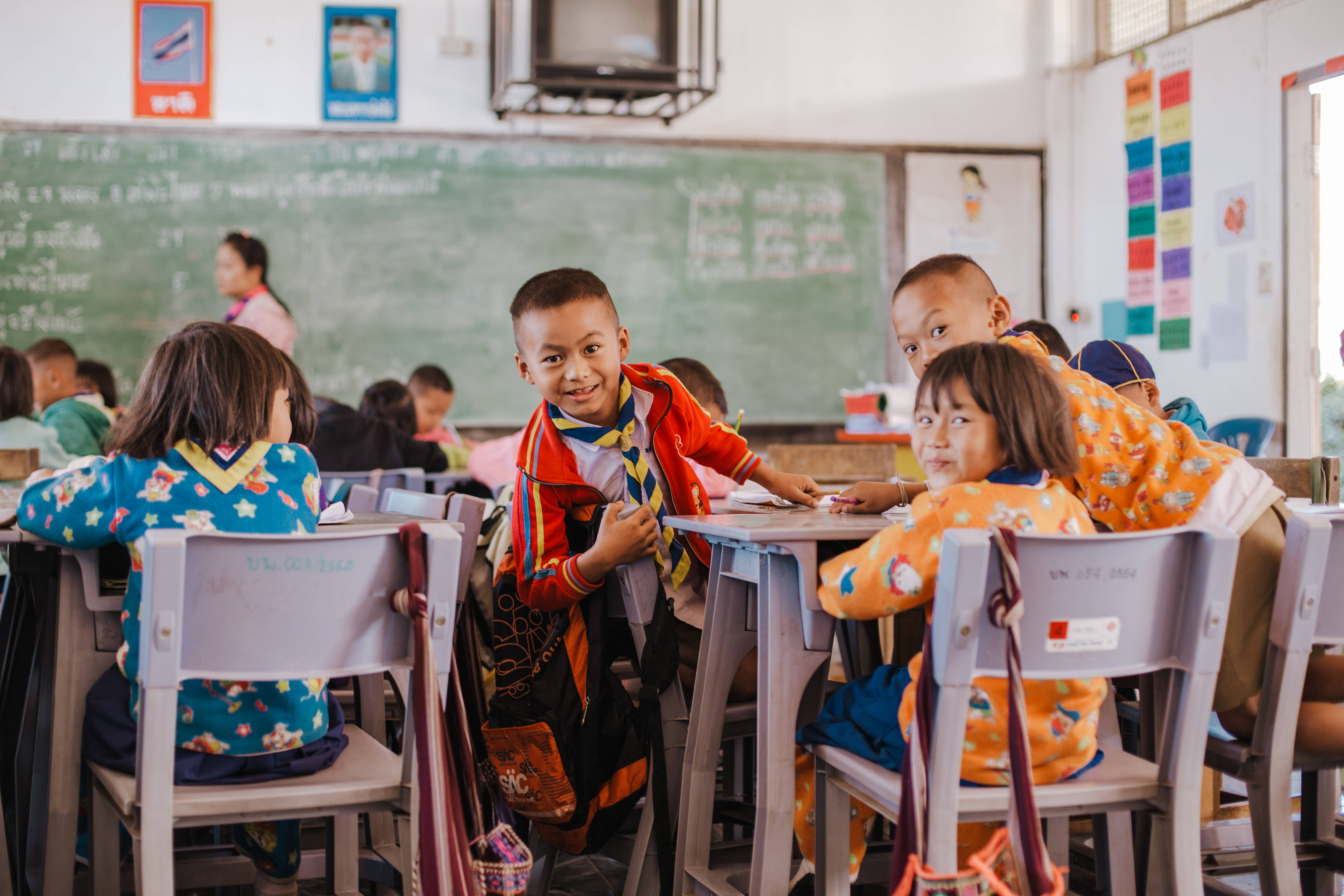 Educational 桌布 壁紙 — 一個穿著紅色制服的快樂小男孩對著鏡頭微笑，而同學們則坐在有綠色黑板的明亮教室的書桌旁。