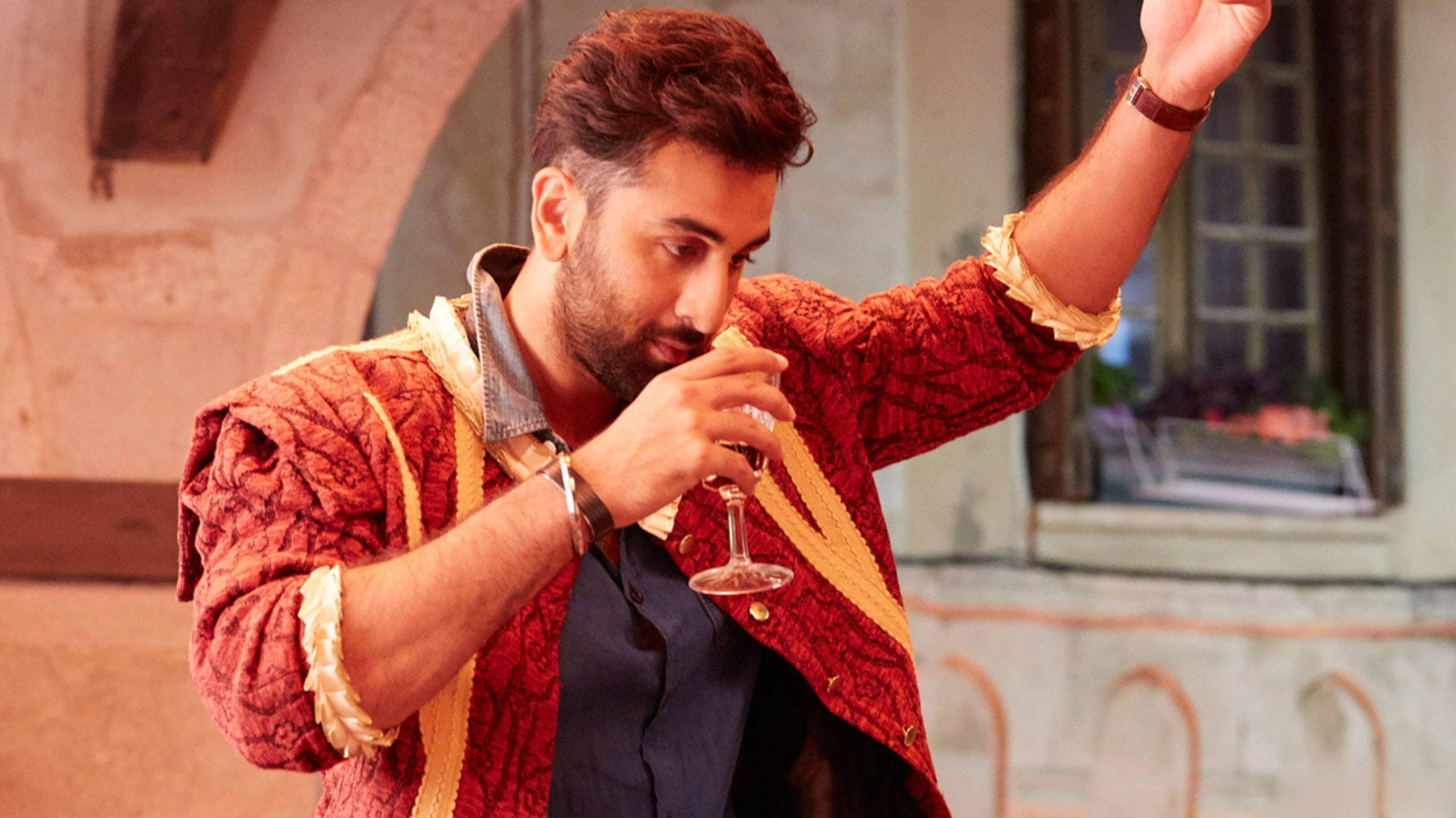 Ranbir Kapoor 桌布 壁紙 — 一個穿著紅色圖案外套的男人在陽光明媚的庭院裡一邊啜飲葡萄酒，一邊用另一隻手做手勢。