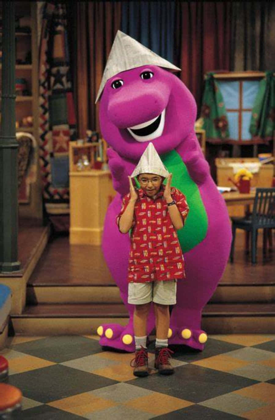 barney 桌布 壁紙 — 一個穿著紅色圖案襯衫和紙帽的孩子站在一個巨大的紫色巴尼恐龍吉祥物旁邊，吉祥物戴著相配的帽子，在色彩繽紛的室內場景中。