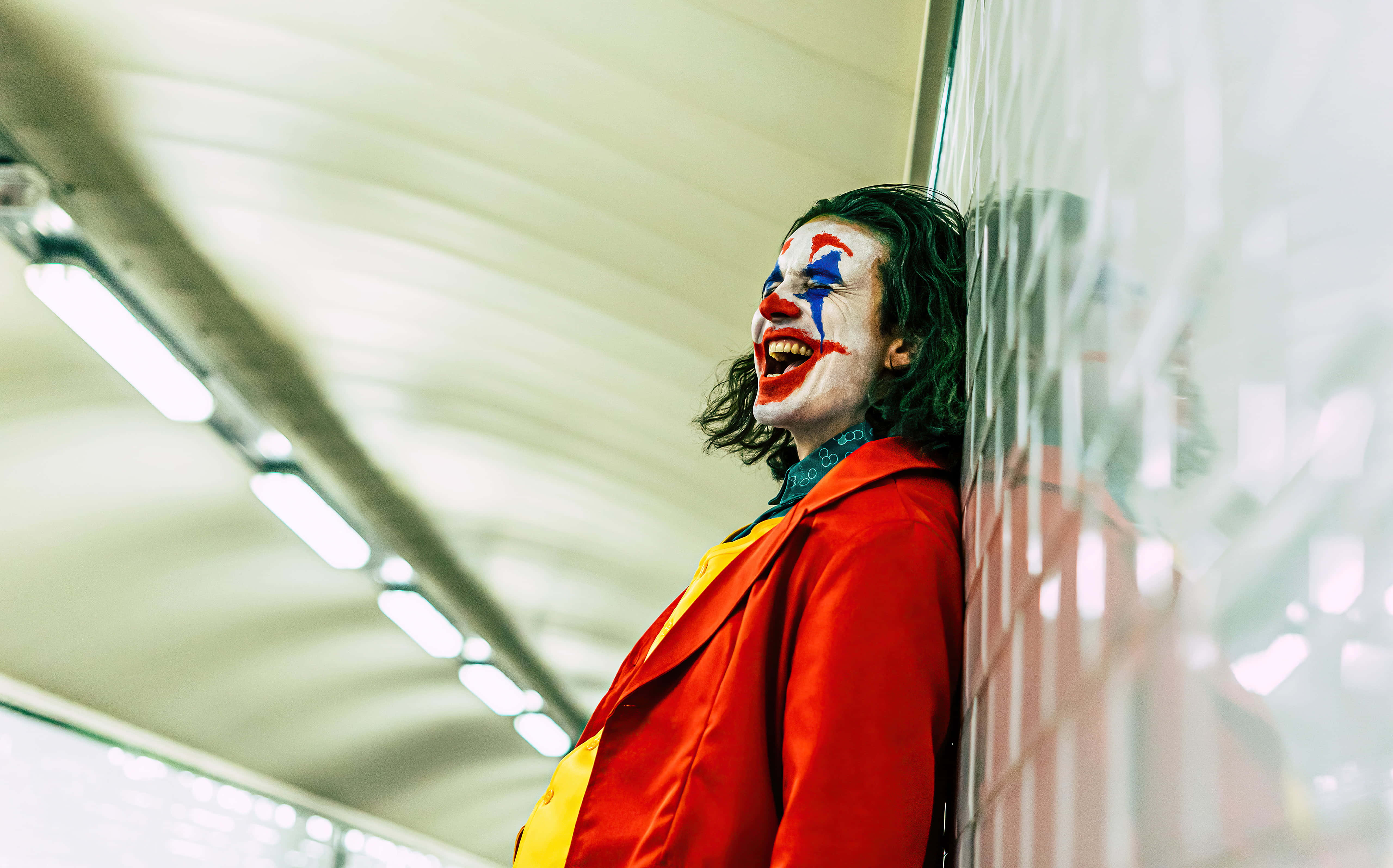 Joker Laughing 桌布 壁紙 — 一個穿著紅色外套、化著小丑妝的人，在光線明亮的地鐵站裡靠著瓷磚牆狂笑。
