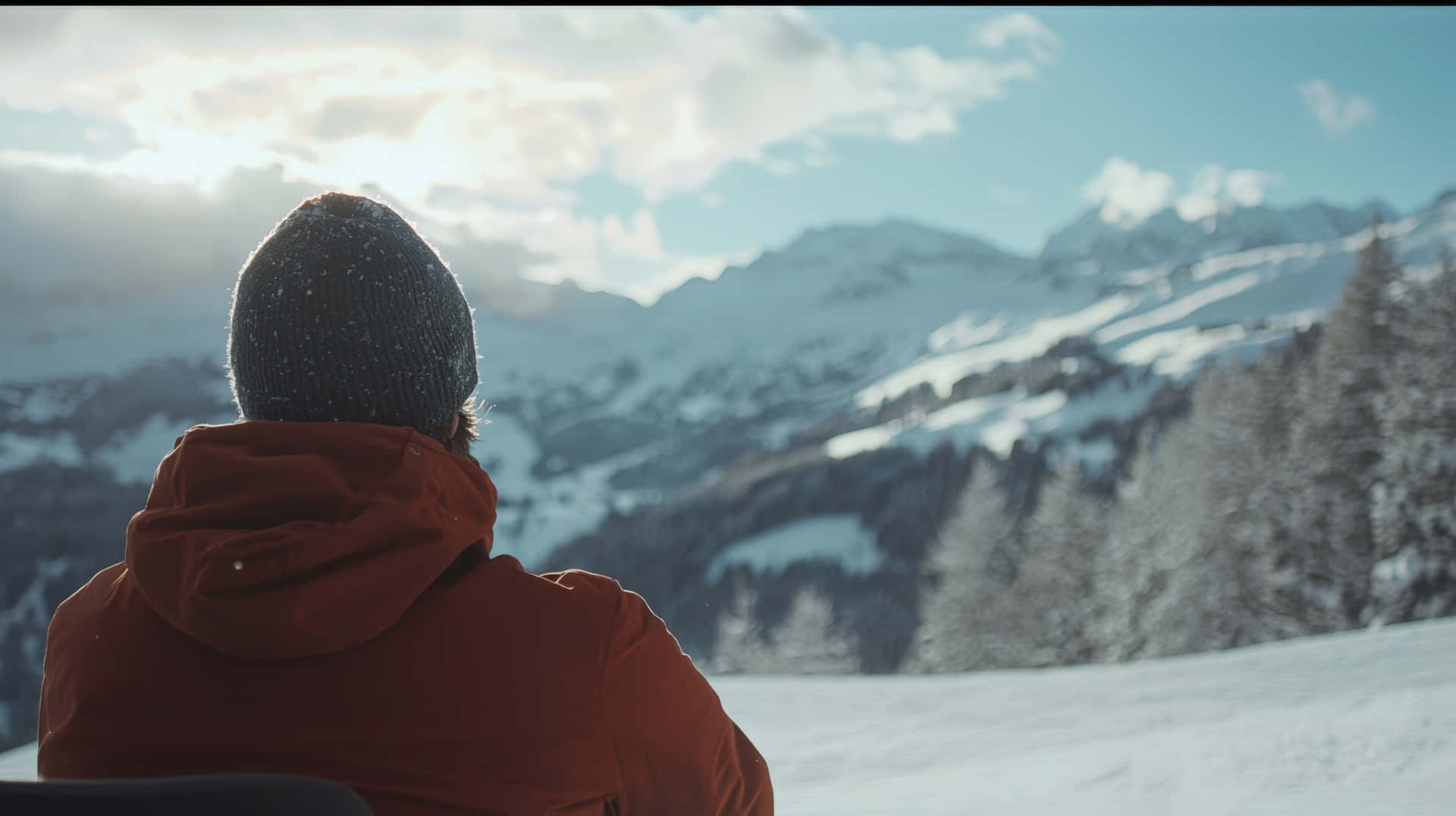 Man in winter clothing admiring the scenic snowy mountain landscape at sunrise 壁紙 — 一個穿著紅色外套和深色毛帽的人坐在雪山上，面向明亮的天空，天空中有散落的雲。