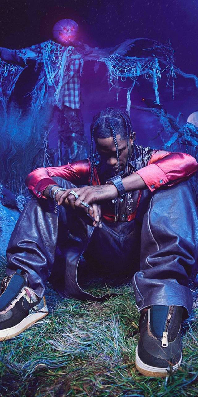 Travis Scott Aesthetic 桌布 壁紙 — 一個穿著紅色外套和黑色皮褲的男人坐在草地上，周圍是詭異的紫色燈光，身後有一個發光的稻草人。