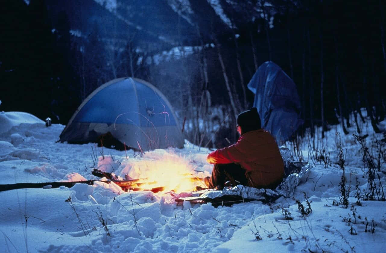 雪 桌布 壁紙 — 一個穿著紅色外套的人坐在雪地森林夜晚中發光的營火旁，附近有兩個帳篷。