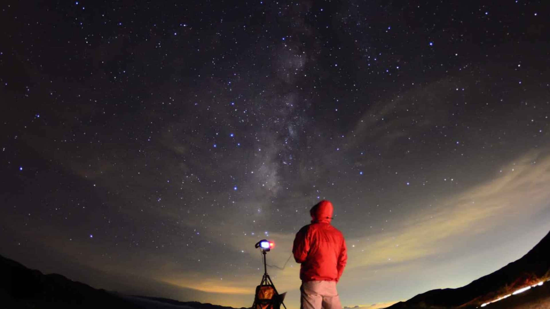 Starry Sky 桌布 壁紙 — 一個穿著紅色外套的人夜晚站在山脊上，仰望星空，銀河在上方閃耀。