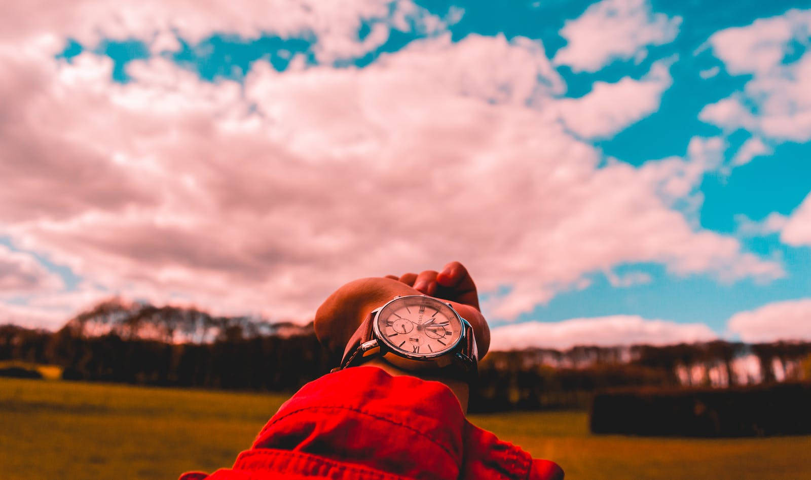Time 桌布 壁紙 — 一個穿著紅色外套的人舉起手錶，背景是充滿活力的粉紅色和藍色天空，有蓬鬆的雲朵。