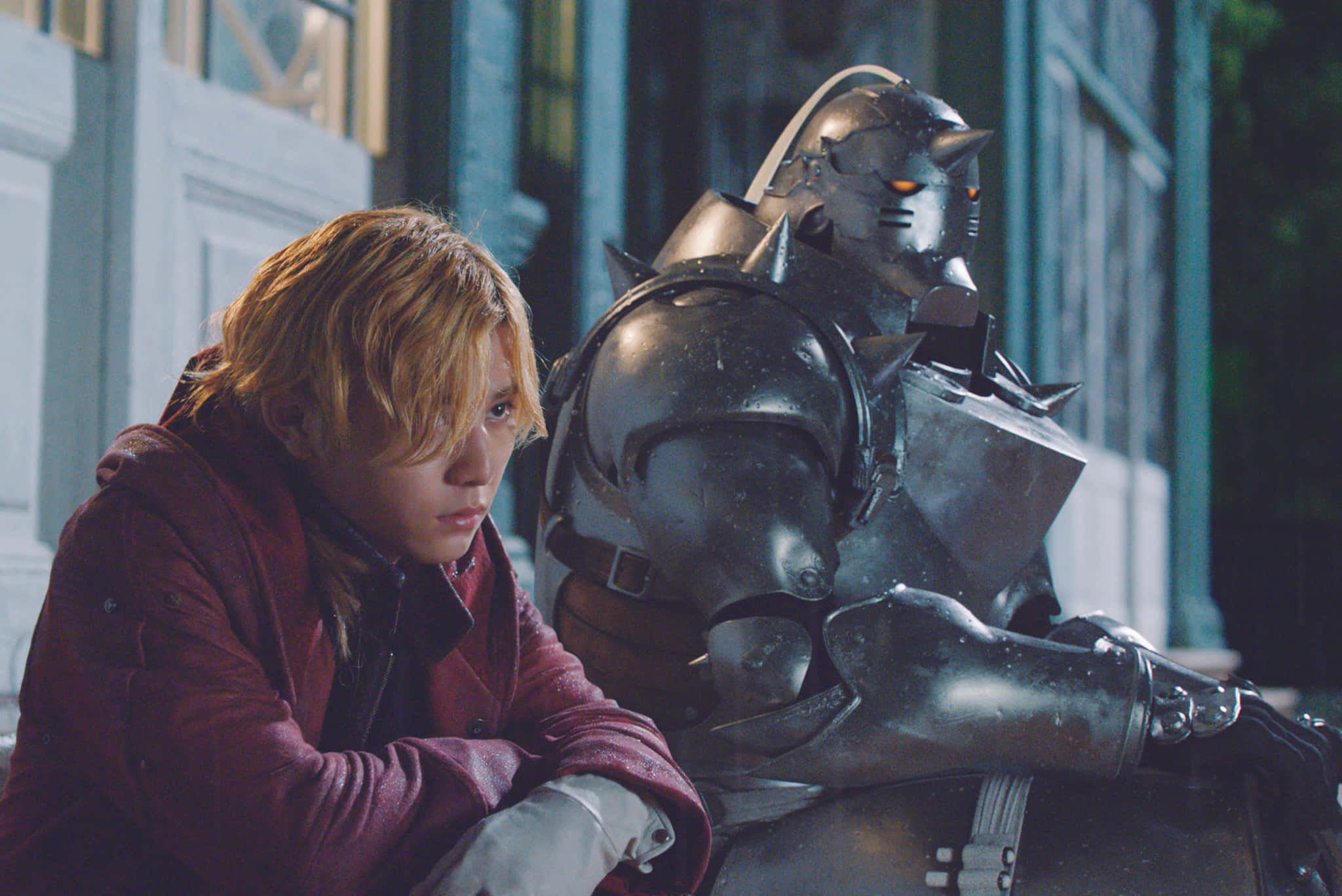Fullmetal Alchemist 桌布 壁紙 — 一個穿著紅色外套的年輕男子坐在一個昏暗的城市小巷裡，旁邊是一個高大的裝甲自動機，有著發光的橙色眼睛。