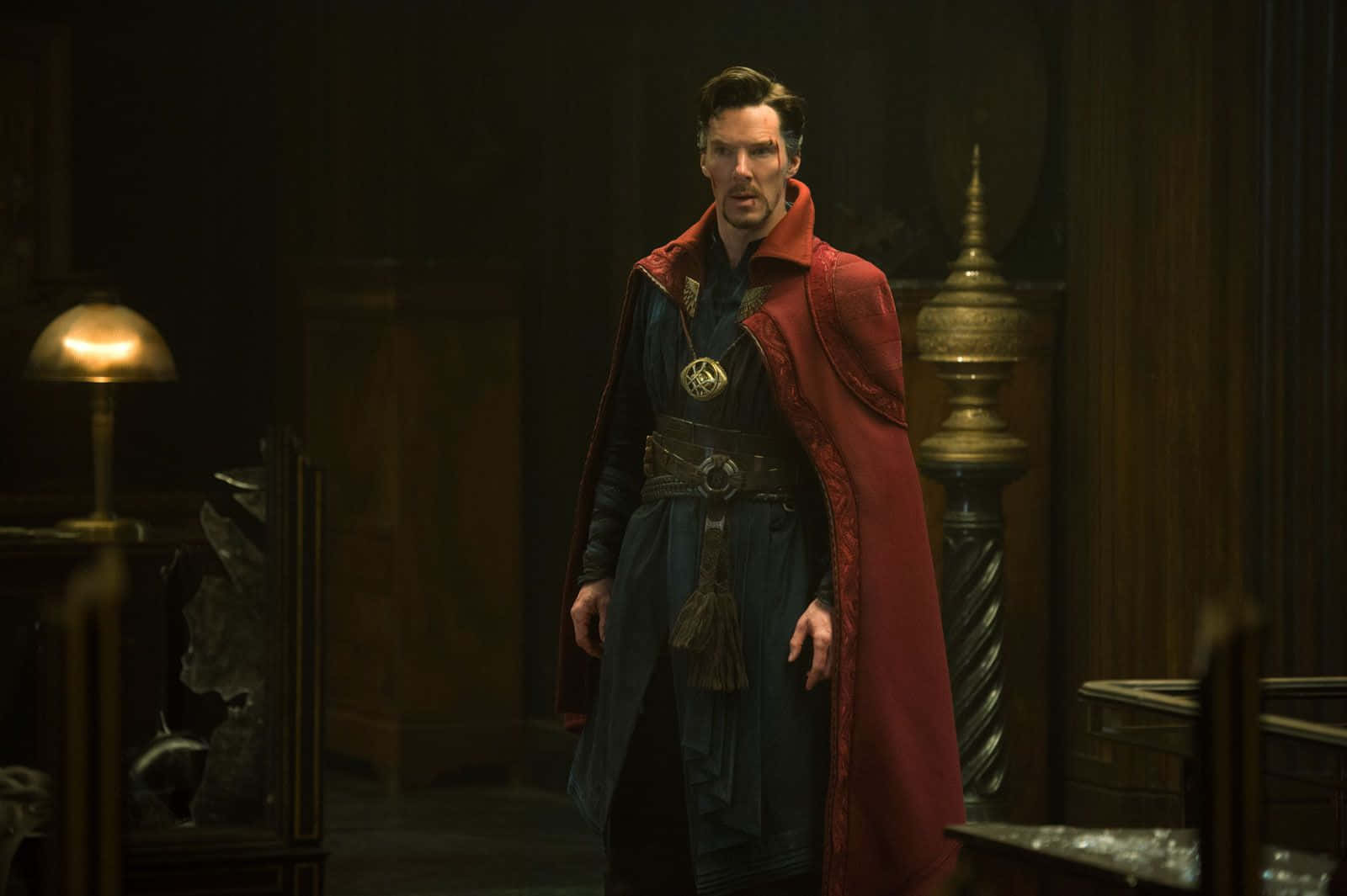 Dr Strange 桌布 壁紙 — 一個穿著紅色斗篷和深色長袍的男人站在一個光線昏暗、裝飾華麗的房間裡，房間裡有復古家具和溫暖的燈光。