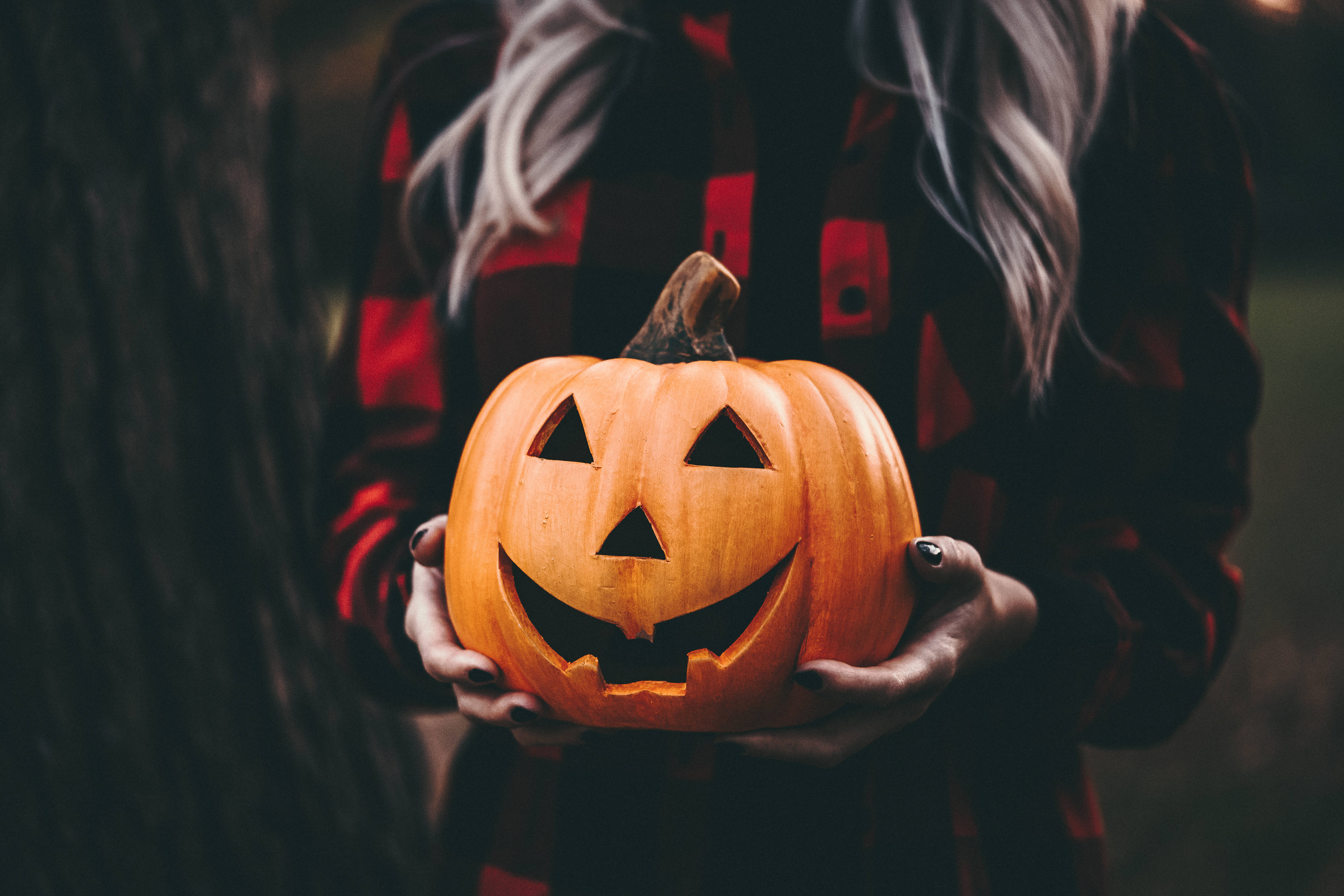 Most Scary 桌布 壁紙 — 一個穿著紅色格紋襯衫的人在黑暗森林背景下拿著一個雕刻了發光面孔的橙色南瓜。