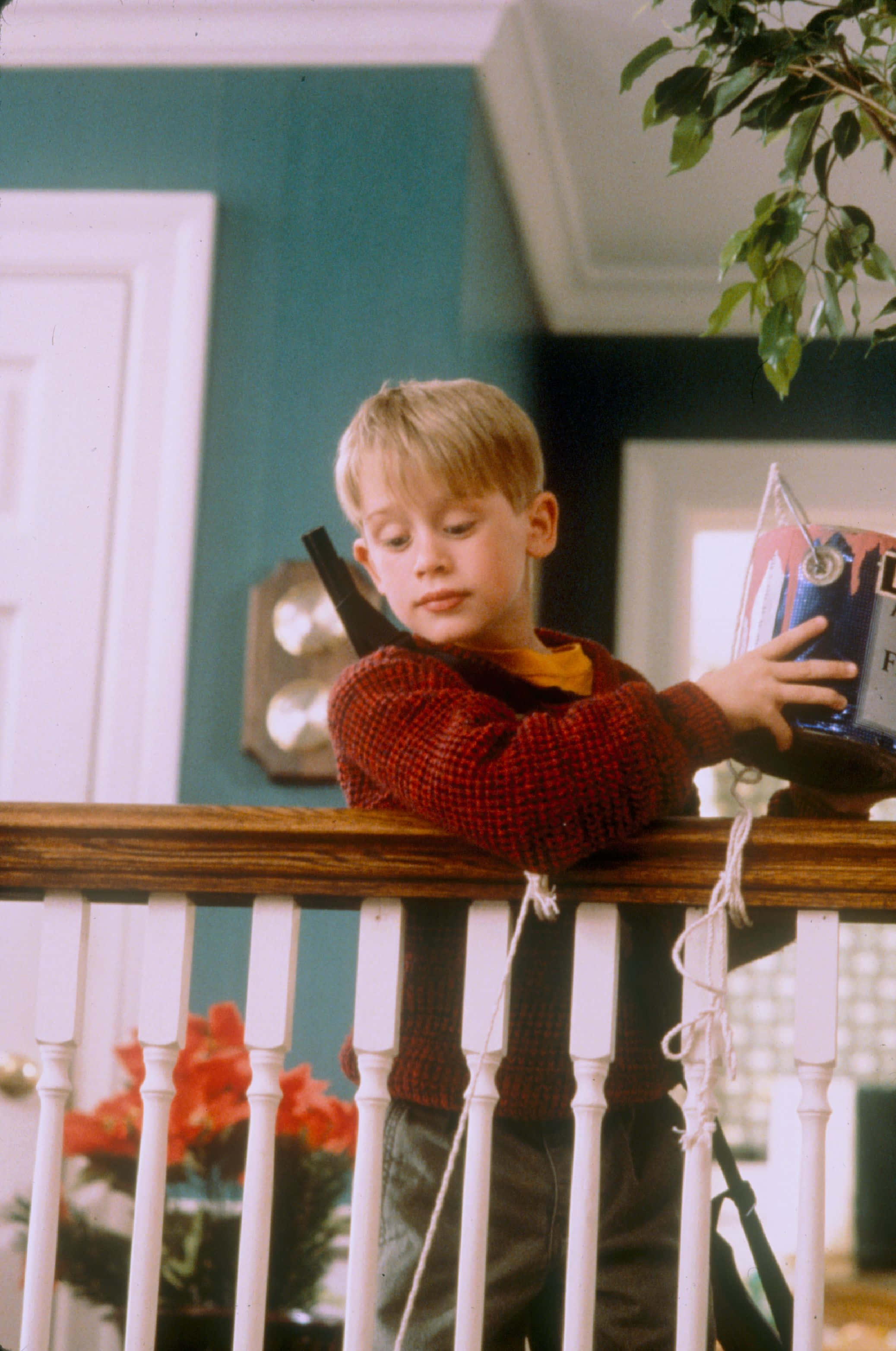 Home Alone 桌布 壁紙 — 一個穿著紅色毛衣的小男孩靠在白色門廊欄杆上，拿著一份包裝好的禮物，表情若有所思。