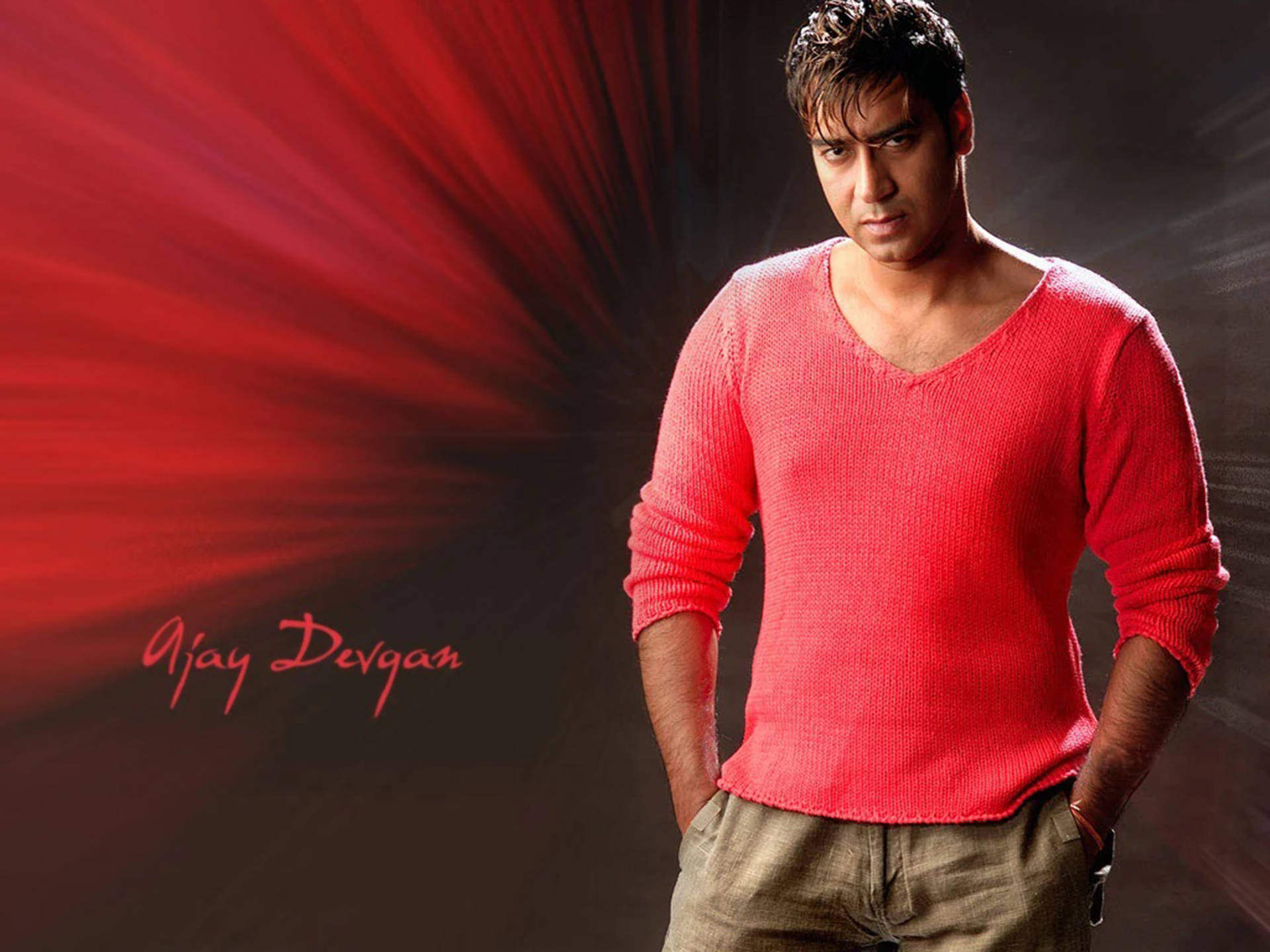 ajay devgn 桌布 壁紙 — 一個穿著紅色毛衣的男人自信地站在戲劇性的紅色放射背景前，他的名字用紅色書寫。