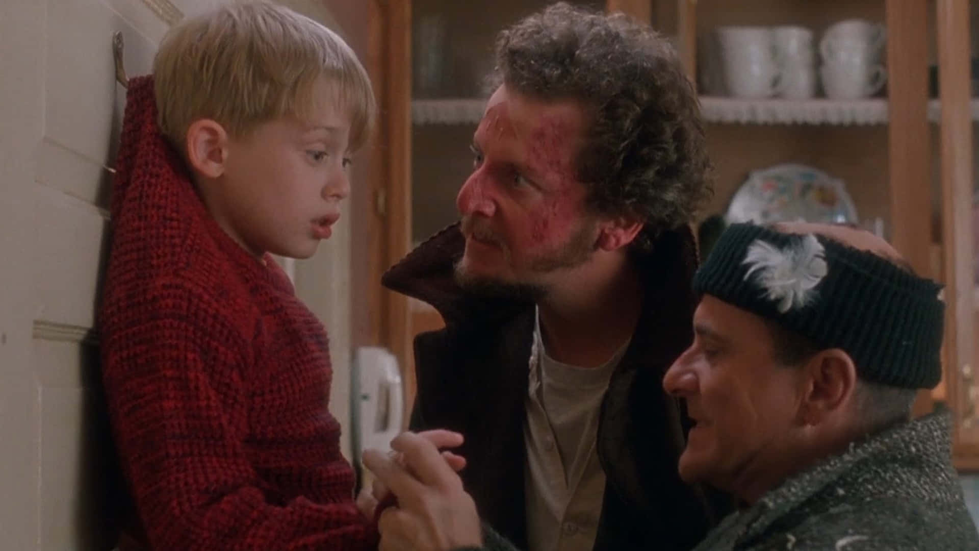 Home Alone 桌布 壁紙 — 一個穿著紅色毛衣的男孩靠牆站著，兩個受傷的男人在廚房裡看著他。