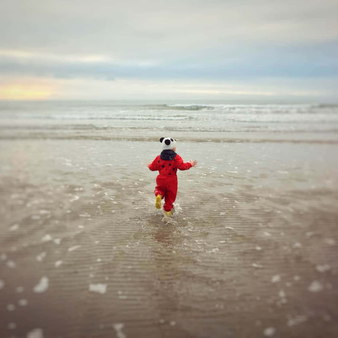 背景 桌布 壁紙 — 一個穿著紅色瓢蟲套裝的幼兒在日落時快樂地跑進海浪中。