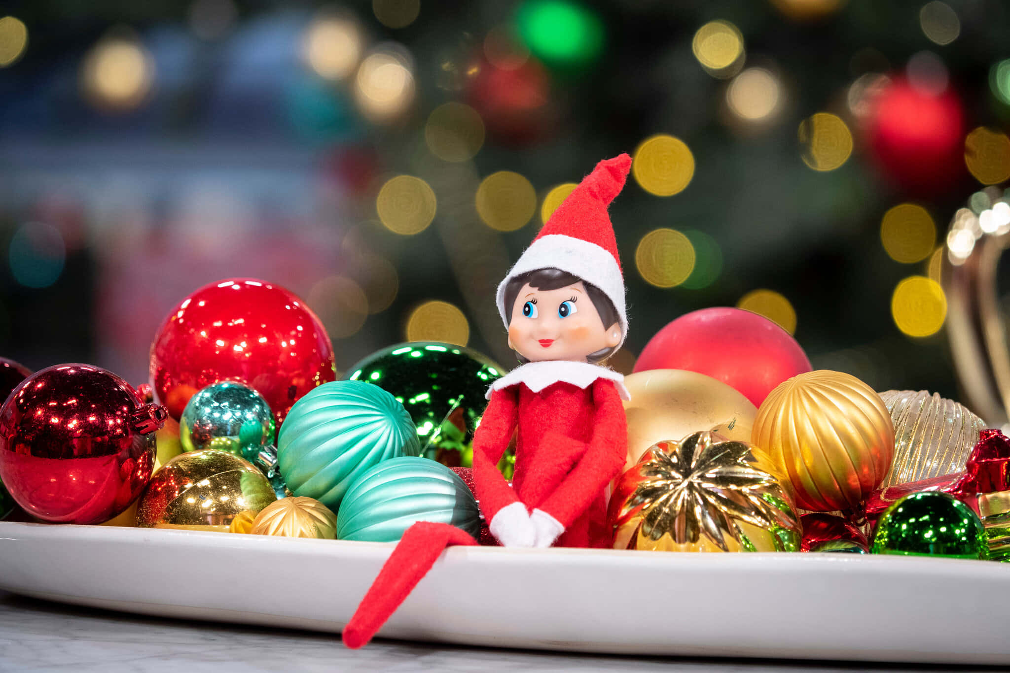 聖誕節 桌布 壁紙 — 一個穿著紅色衣服的快樂精靈公仔坐在白色盤子上，周圍是閃亮的紅色、綠色和金色聖誕裝飾，背景有散景燈光。