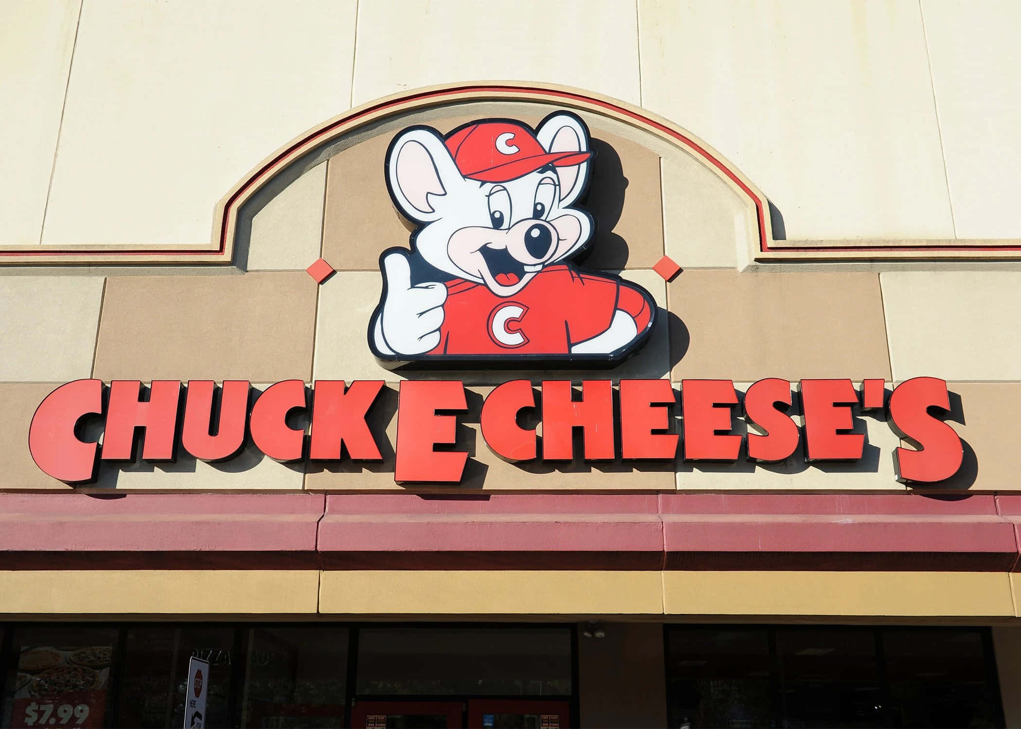 查克芝士 桌布 壁紙 — 一個穿著紅色衣服的開心 Chuck E Cheese 吉祥物，在米色建築物外牆上，於粗體紅色招牌上方豎起大拇指。
