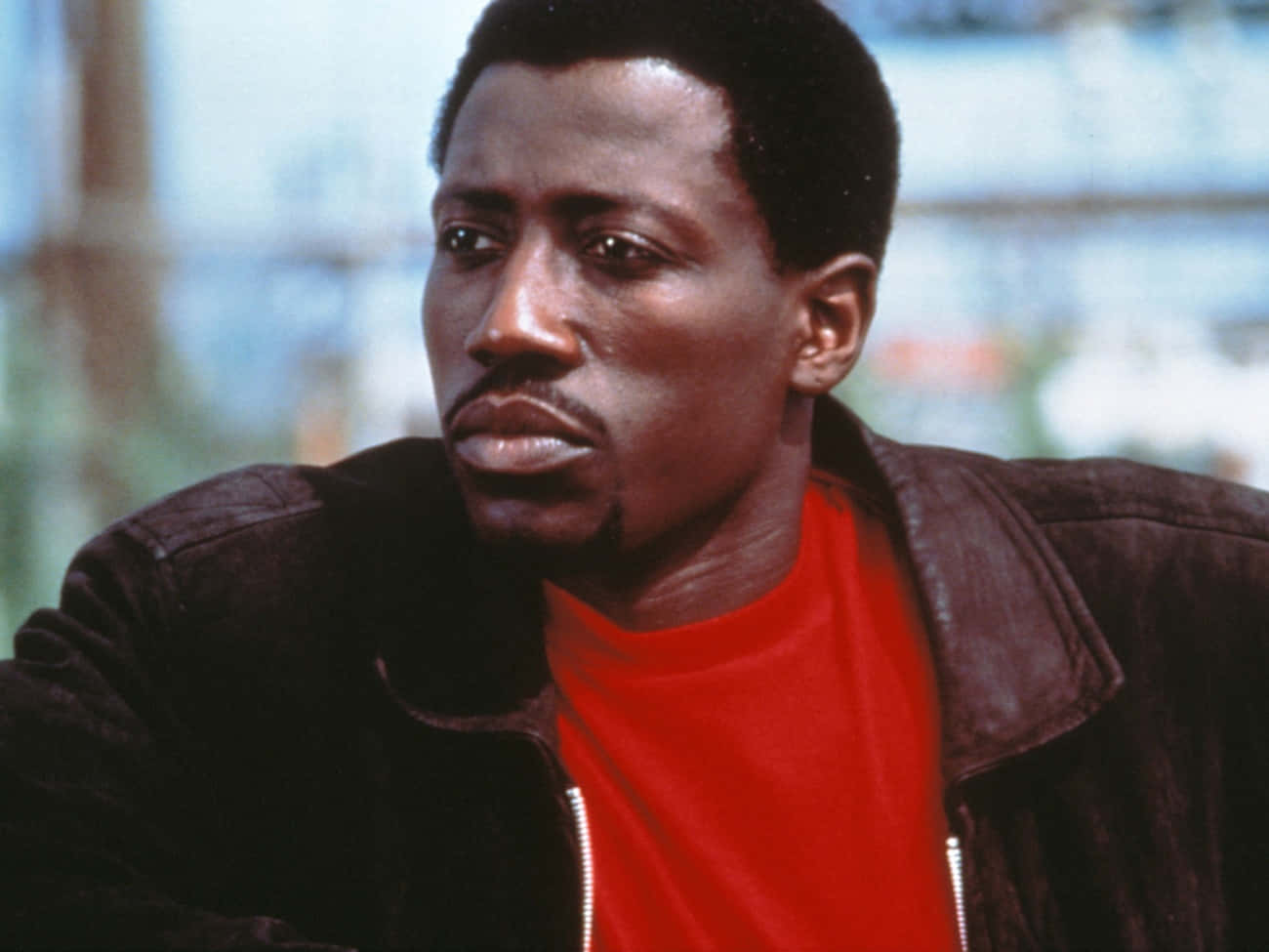 Wesley Snipes 桌布 壁紙 — 一個穿著紅色襯衫和深色外套的男人，在模糊的城市背景下，帶著嚴肅的表情沉思地凝視著。