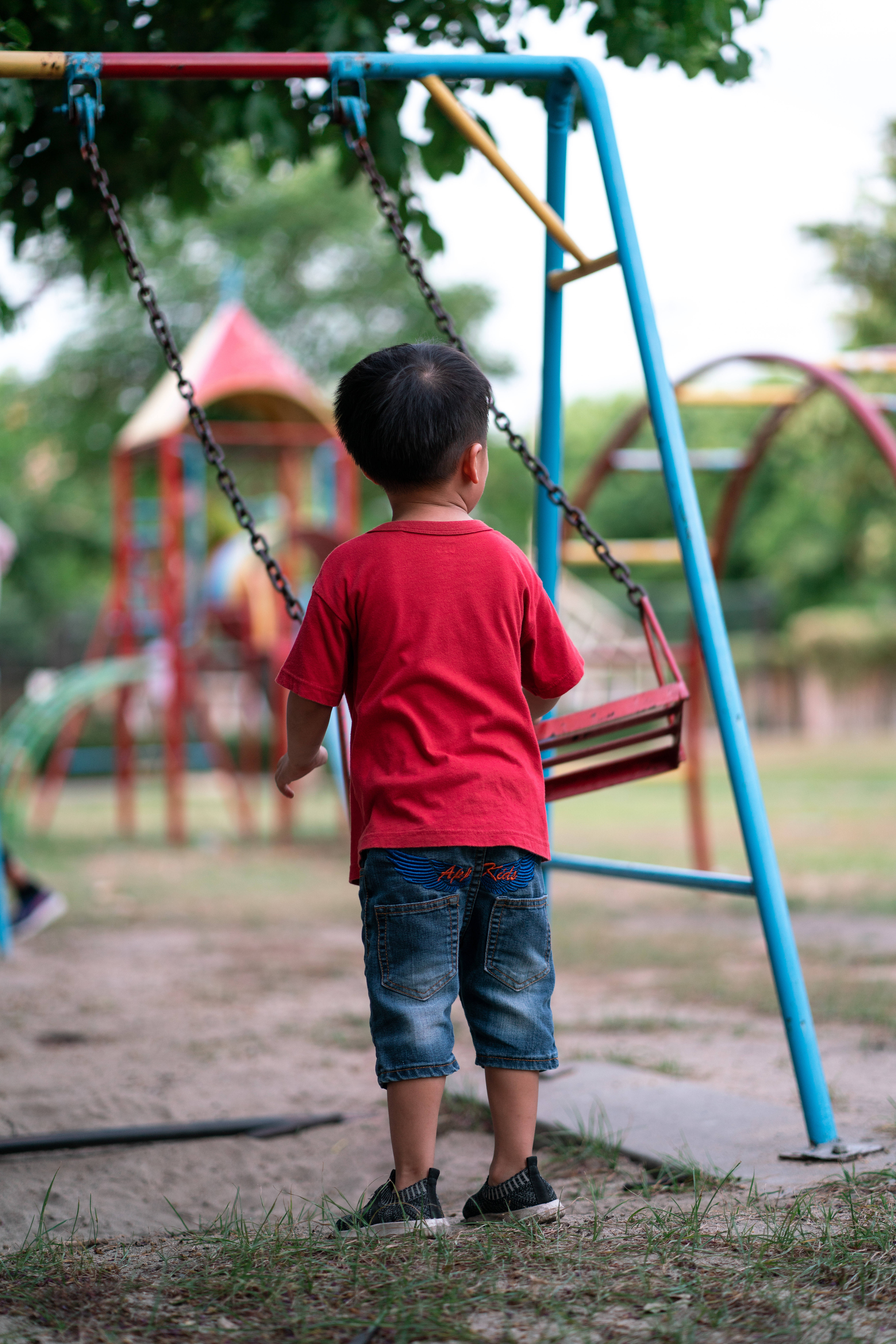 Playground 桌布 壁紙 — 一個穿著紅色襯衫的小男孩獨自站在公園裡一個色彩繽紛的鞦韆旁，周圍有綠樹和柔和的光線。