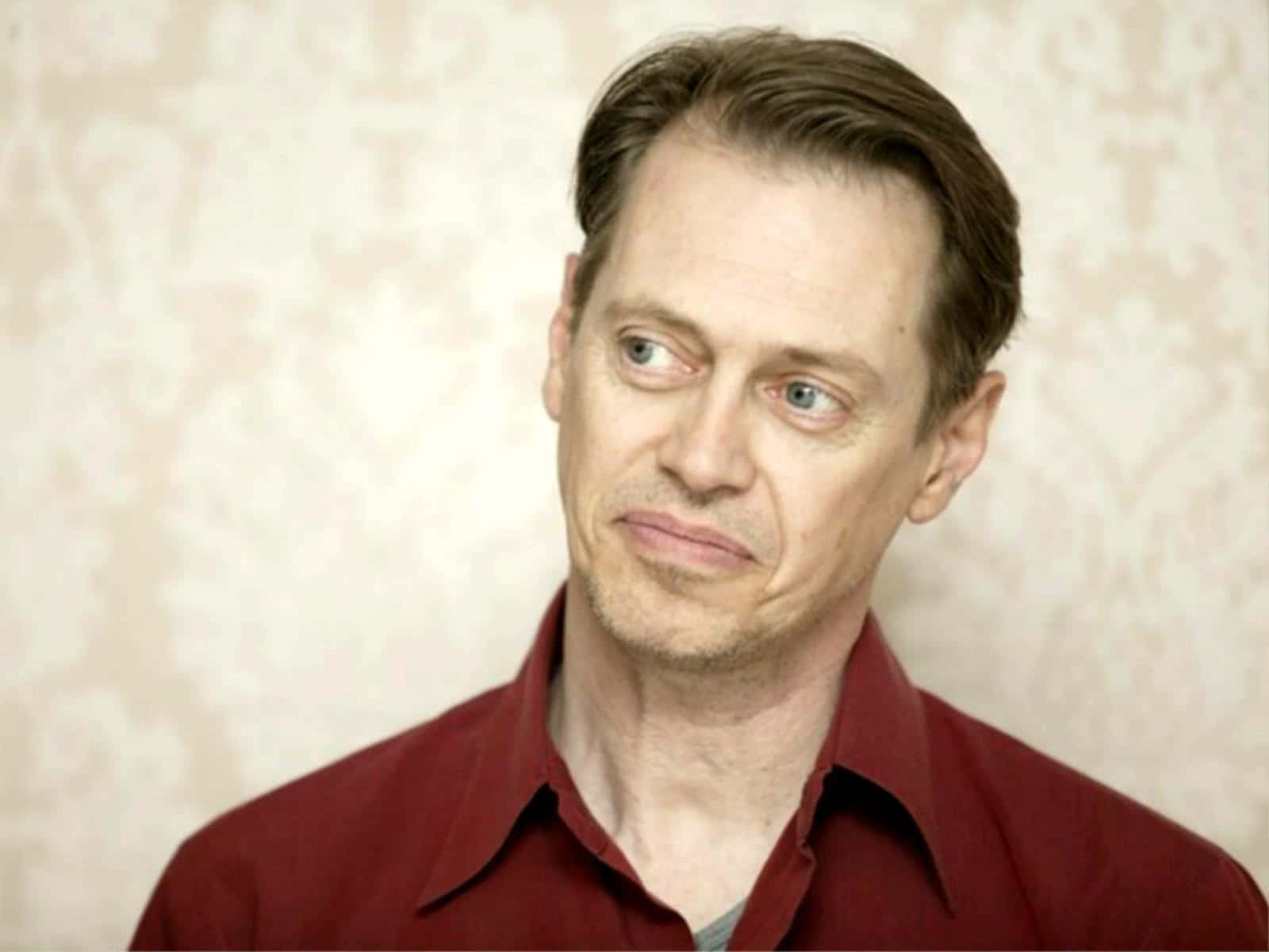 steve buscemi 桌布 壁紙 — 一個穿著紅色襯衫的男人，在帶有圖案的米色壁紙背景下，側面露出懷疑的表情。