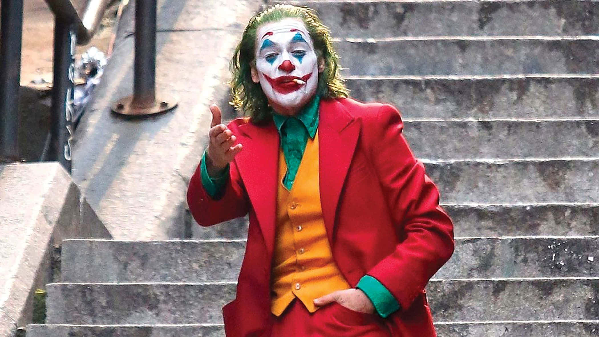 小丑 桌布 壁紙 — 一個穿著紅色西裝和小丑妝容的男人站在混凝土樓梯上，帶著狂躁的笑容指著。