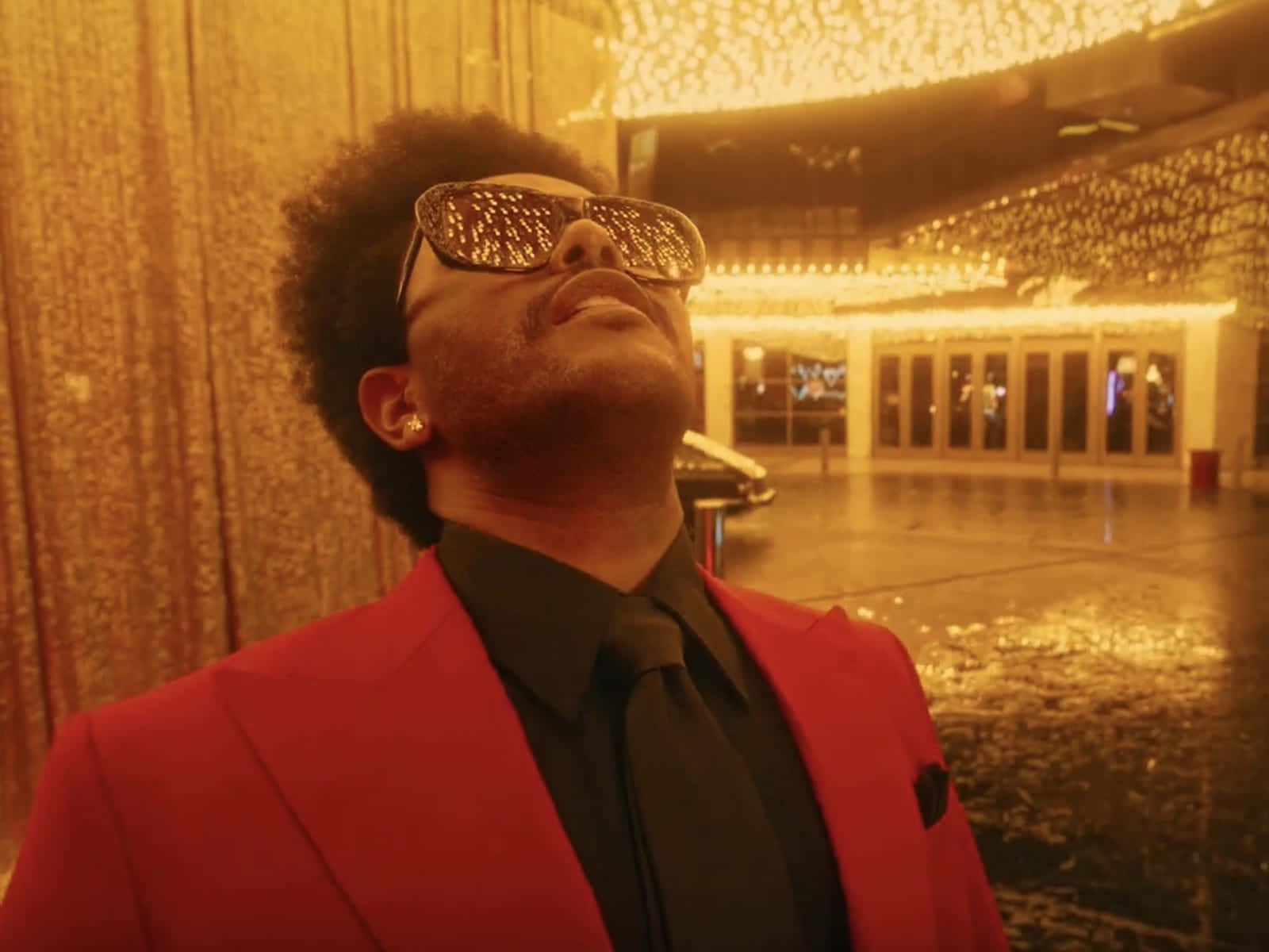 Don Toliver 桌布 壁紙 — 一個穿著紅色西裝和閃亮太陽眼鏡的男人在潮濕的城市庭院中，在金色串燈下向後仰頭。