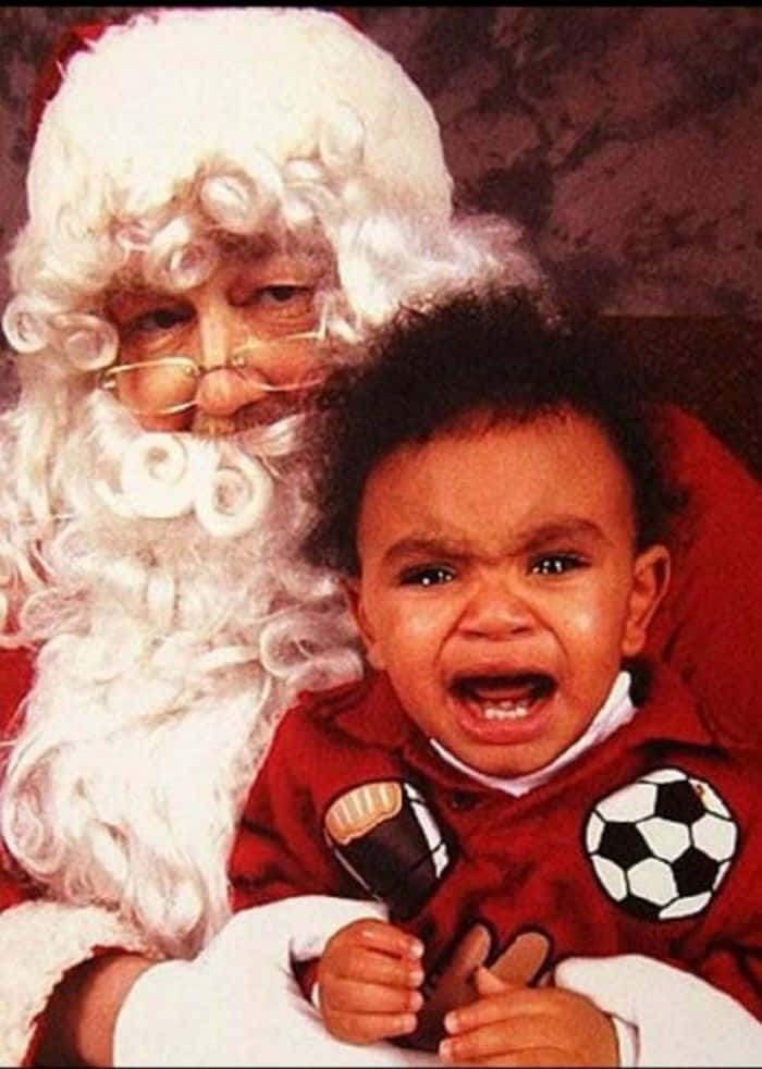 Santa Claus 桌布 壁紙 — 一個穿著紅色足球主題服裝的哭泣嬰兒緊握著麥克風，同時被微笑的聖誕老人抱著。