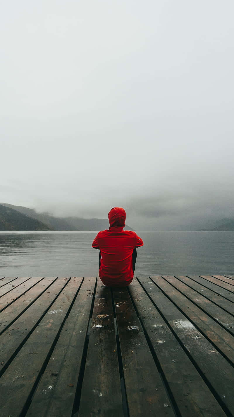 Alone 桌布 壁紙 — 一個穿著紅色連帽外套的人獨自坐在一個經風吹日曬的木碼頭上，眺望著一個霧氣瀰漫的湖泊，遠處是霧濛濛的山巒。