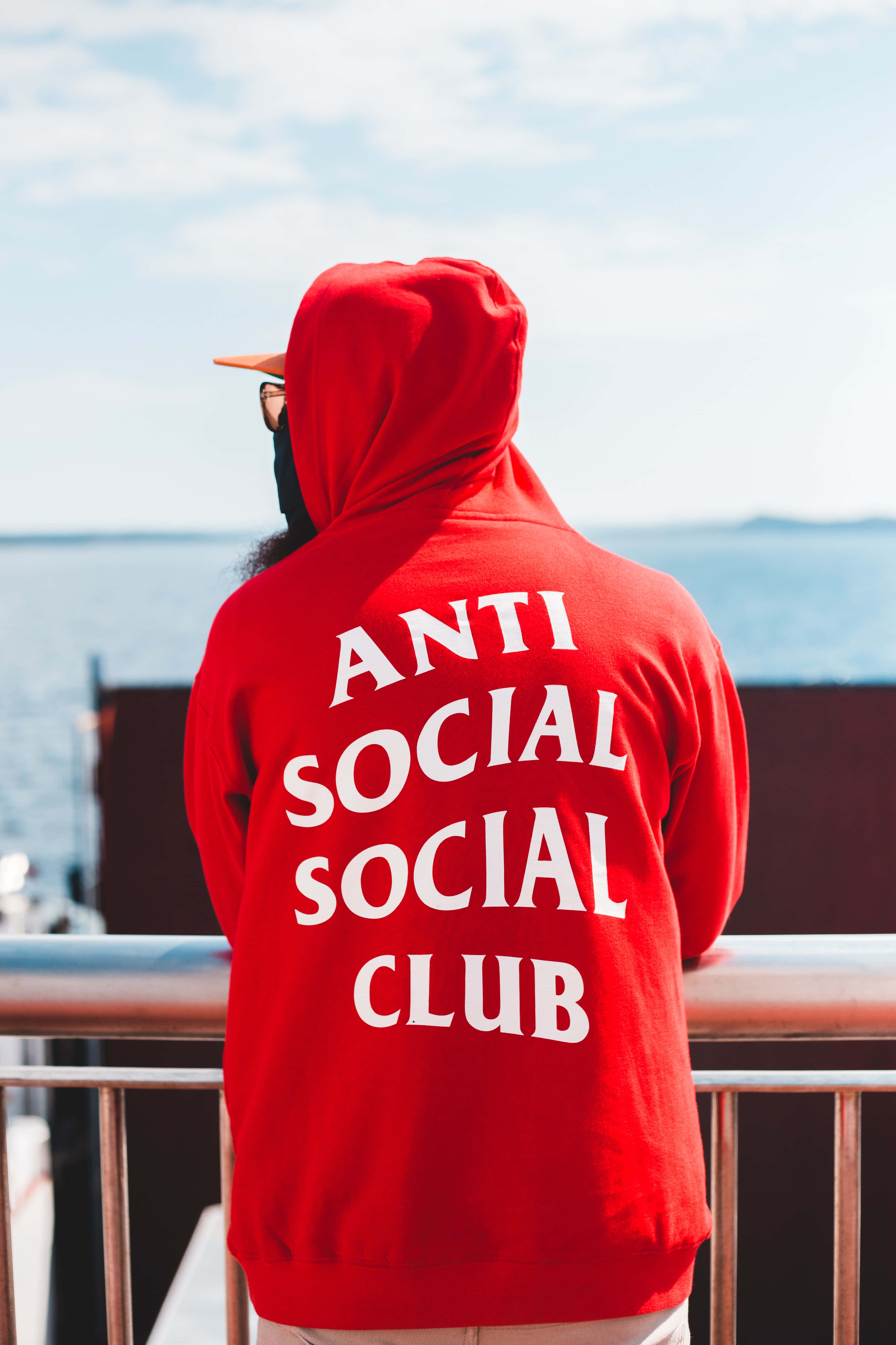 Anti Social Club iPhone 桌布 壁紙 — 一個穿著紅色連帽衫、上面有『ANTI SOCIAL SOCIAL CLUB』字樣的人，站在欄杆上俯瞰著晴朗天空下的海洋。