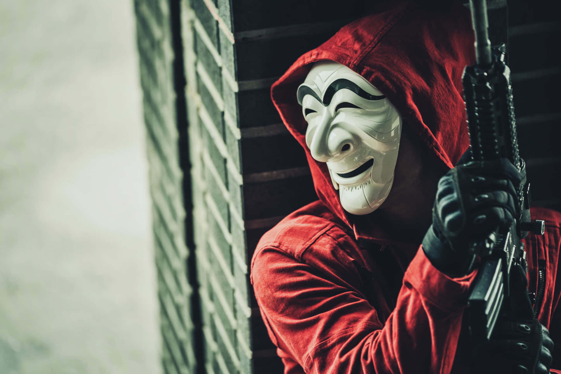 Mask 桌布 壁紙 — 一個穿著紅色連帽衫、戴著白色蓋伊·福克斯面具的人，在昏暗的光線下拿著步槍靠著磚牆。
