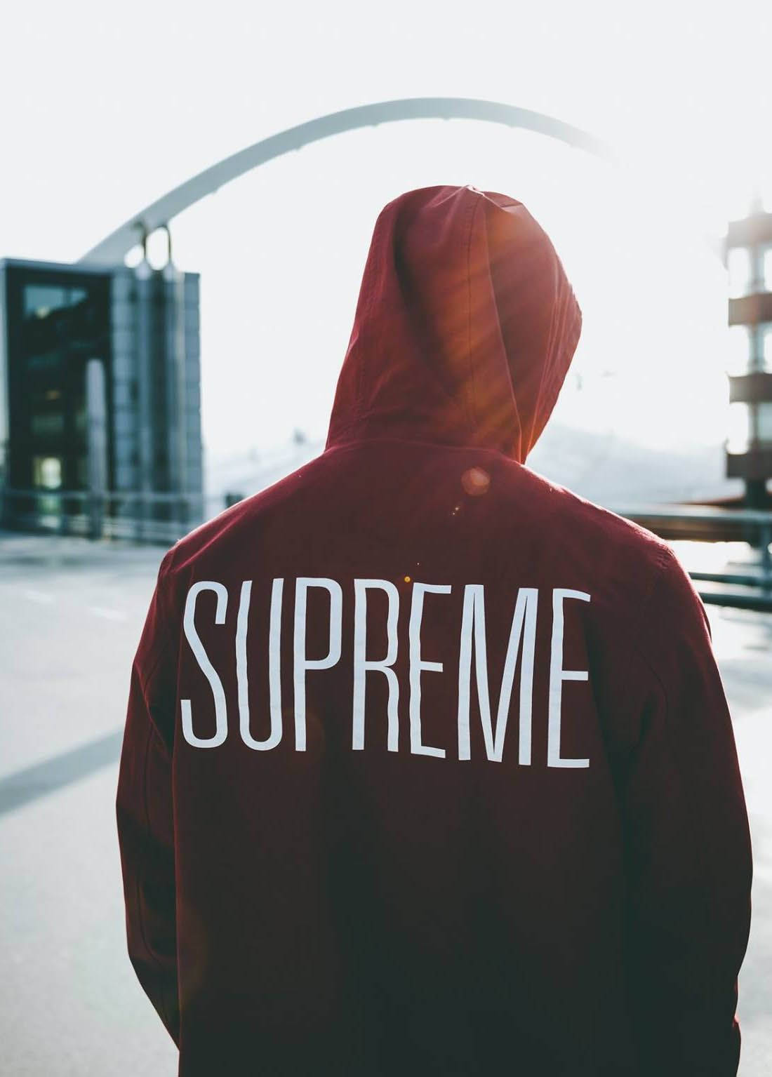 supreme aesthetic 桌布 壁紙 — 一個穿著紅色連帽衫、背面印有「SUPREME」的人背對鏡頭，面向城市環境中的現代拱形建築。