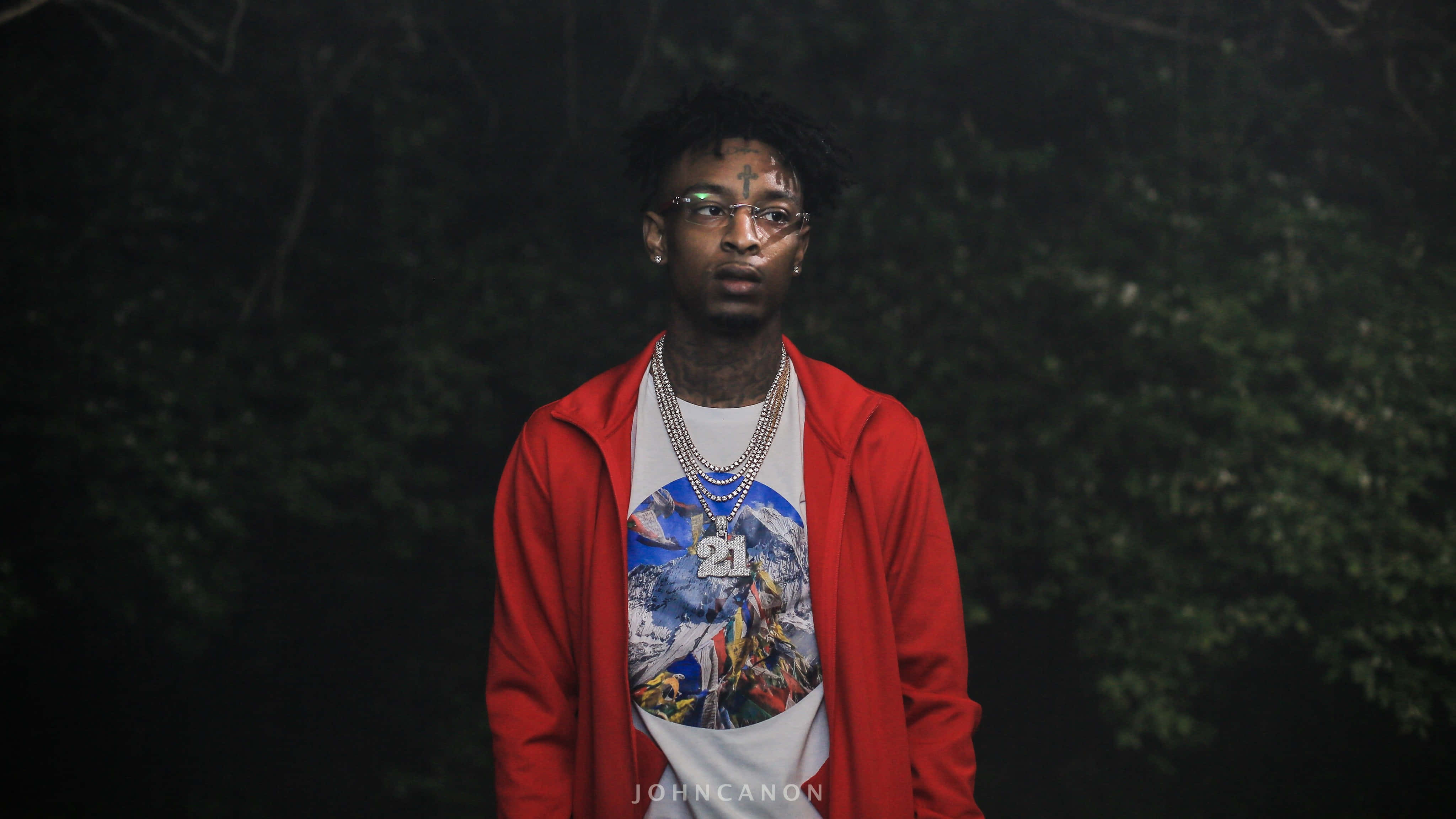 21 Savage 桌布 壁紙 — 一個穿著紅色連帽衫和圖案T恤的年輕人，在夜晚的黑暗森林中自信地站著，眼睛發光。