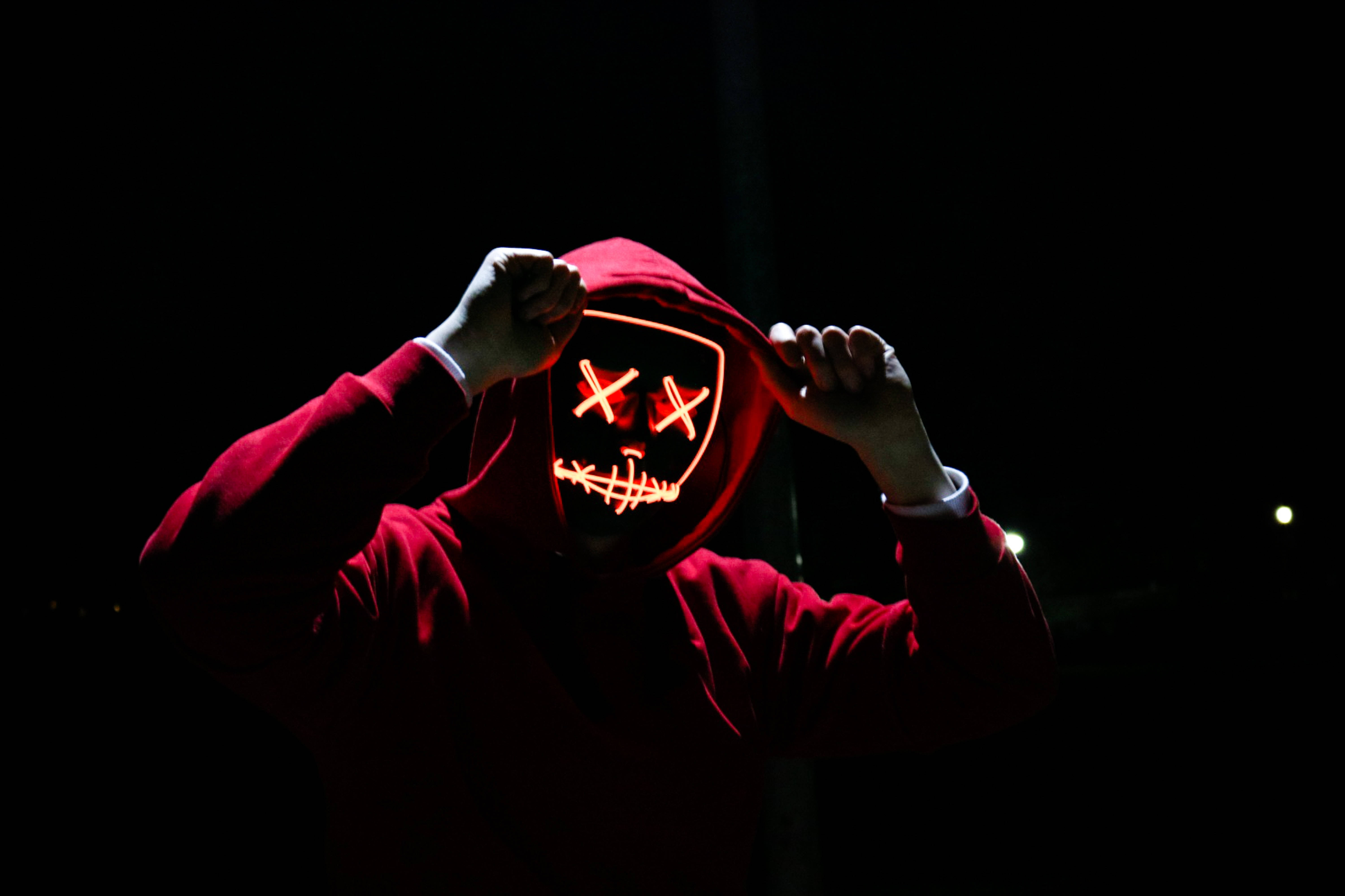 evil smile 桌布 壁紙 — 一個穿著紅色連帽衫的人，在黑暗背景下舉起一個帶有X形眼睛和縫合嘴巴的發光橙色LED面具。