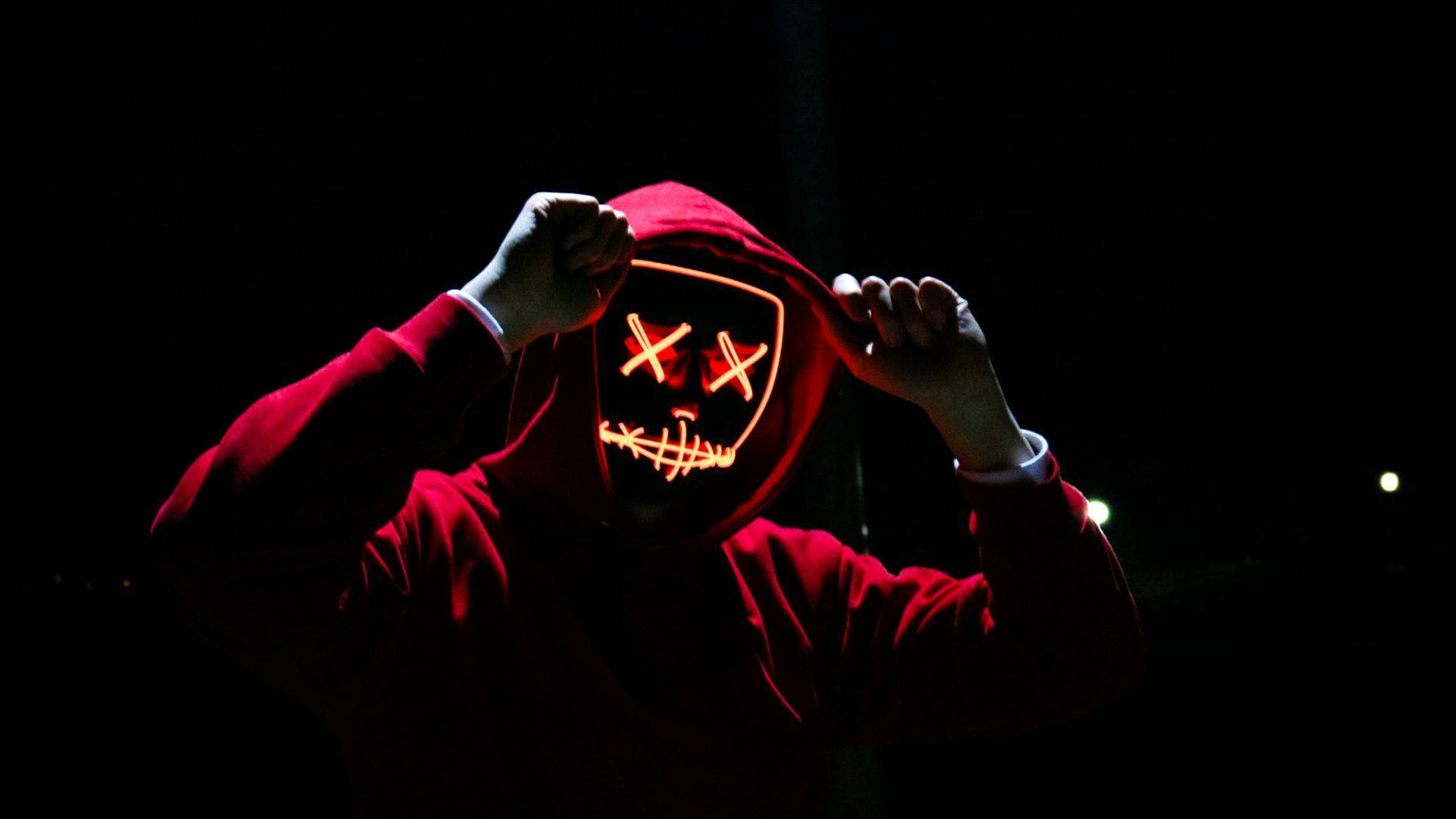 Mask 桌布 壁紙 — 一個穿著紅色連帽衫的人，在黑暗背景前舉起一個發光的LED面具，面具上有X形眼睛和縫合的嘴巴。