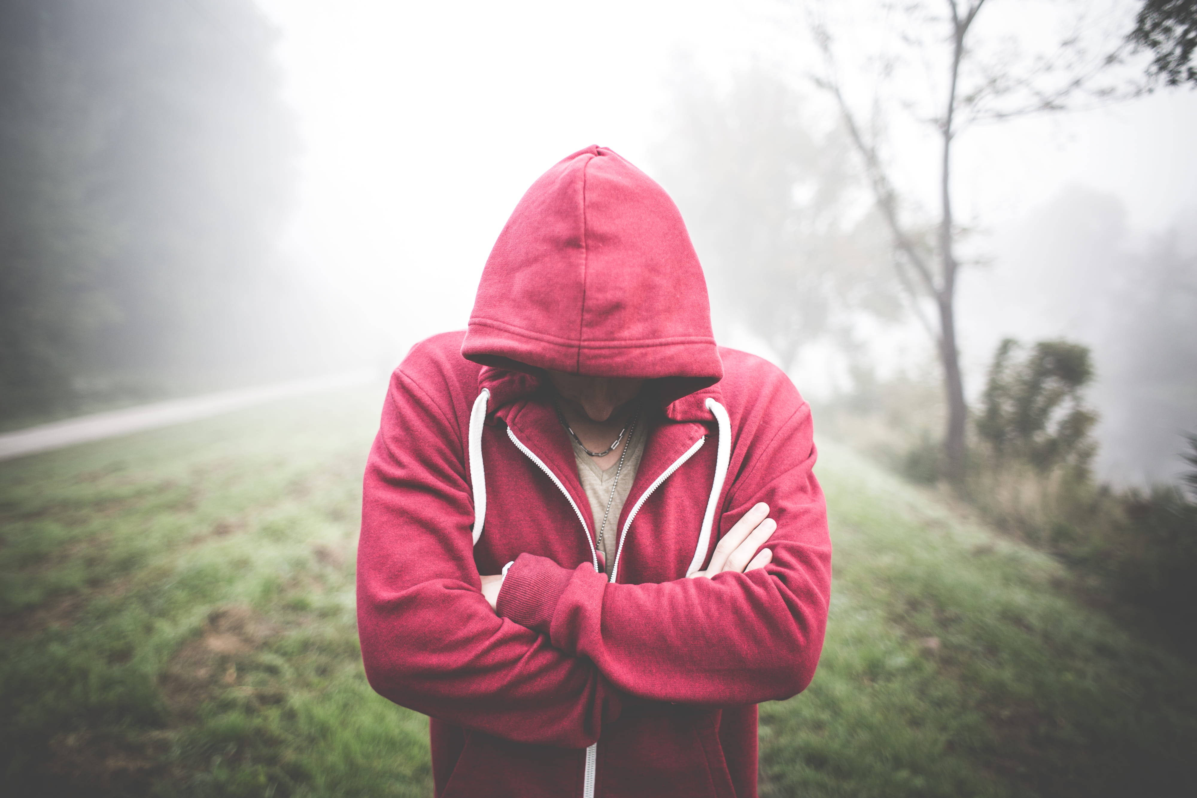 Alone Boy 桌布 壁紙 — 一個穿著紅色連帽衫的人，雙手交叉，低著頭，站在霧氣瀰漫的田野中，周圍是柔和的綠草和光禿禿的樹木。