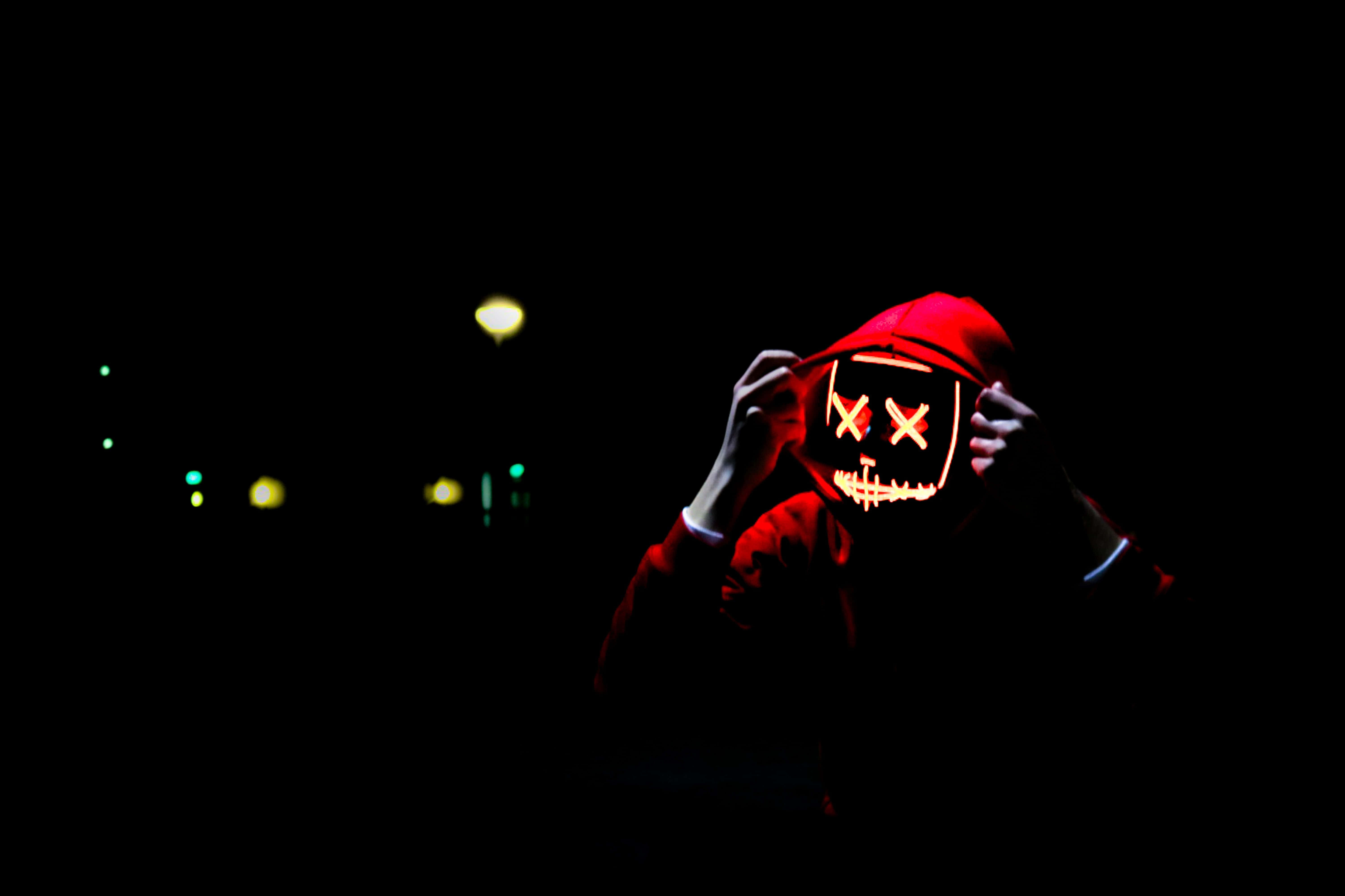 Red Hoodie 桌布 壁紙 — 一個穿著紅色連帽衫的人手持一個發光的骷髏面具，面具上有X形眼睛，背景是漆黑的夜晚和遠處的城市燈光。