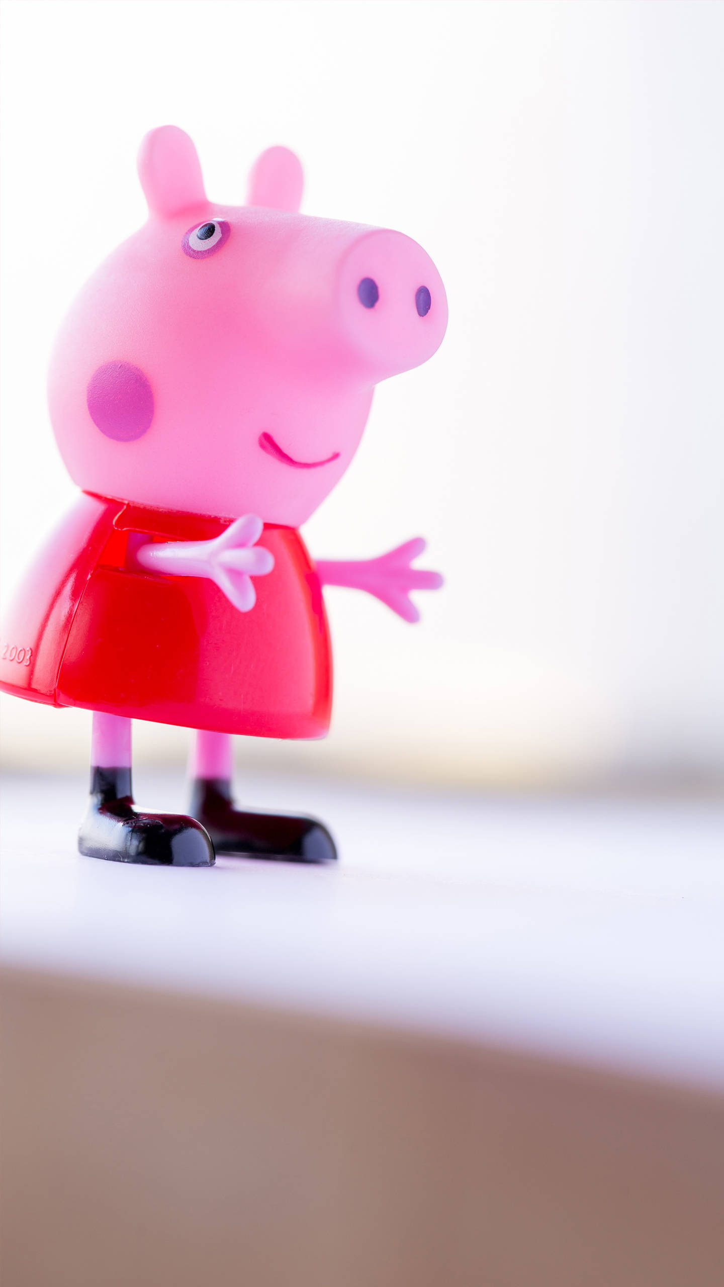 Peppa 桌布 壁紙 — 一個穿著紅色連衣裙的快樂粉紅色塑膠Peppa Pig玩具，站在白色表面上，背景柔和模糊。