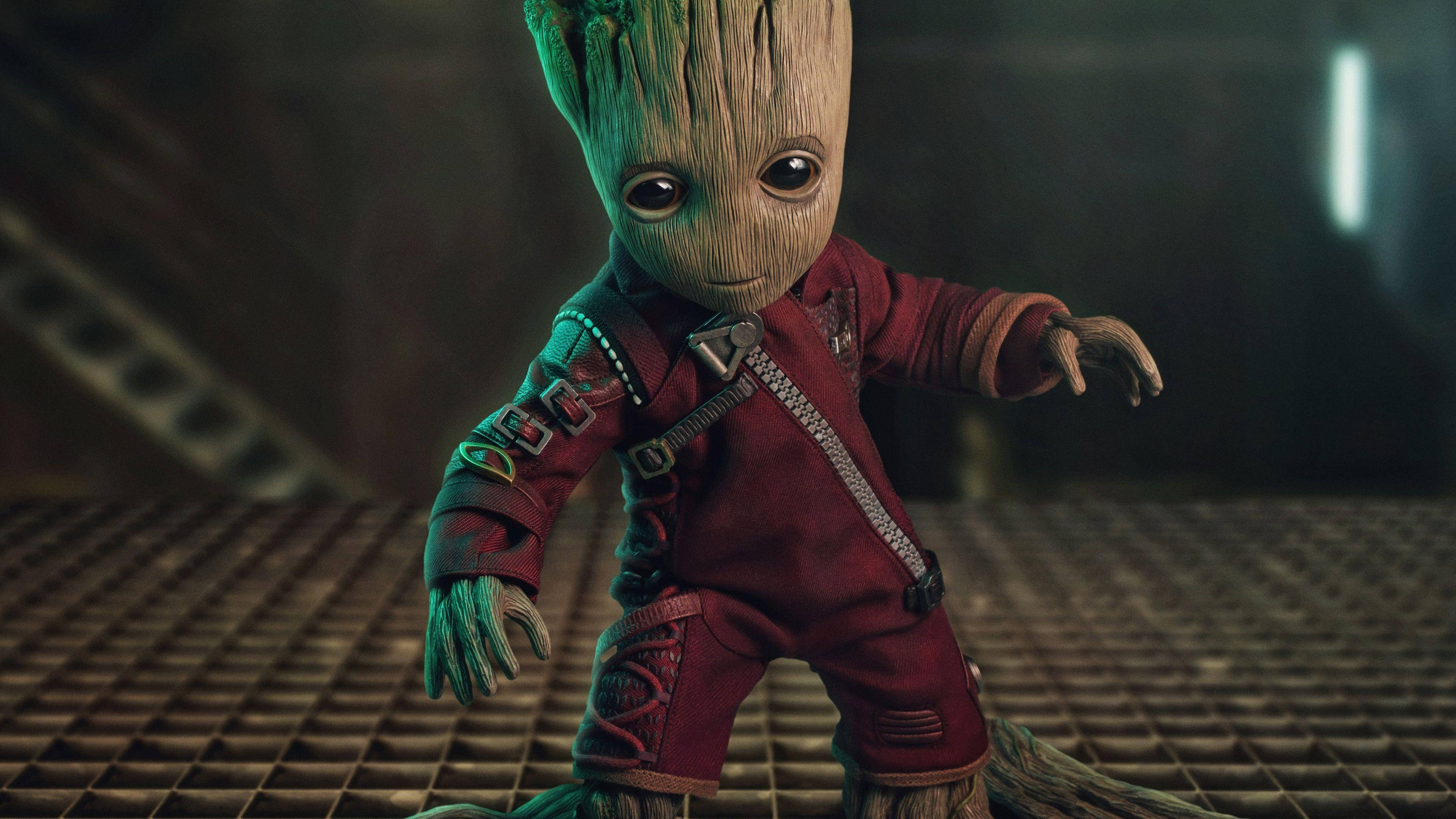 4K Guardians Of The Galaxy 桌布 壁紙 — 一個穿著紅色連身衣的小型樹狀生物站在昏暗工業環境的金屬格柵上。