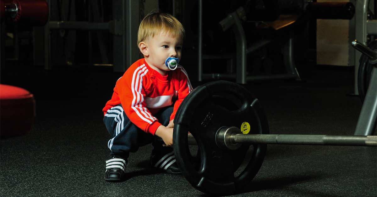Weight Lifting 桌布 壁紙 — 一個穿著紅色運動服的幼兒在昏暗的健身房裡，嘴裡含著奶嘴，握著沉重的槓鈴。