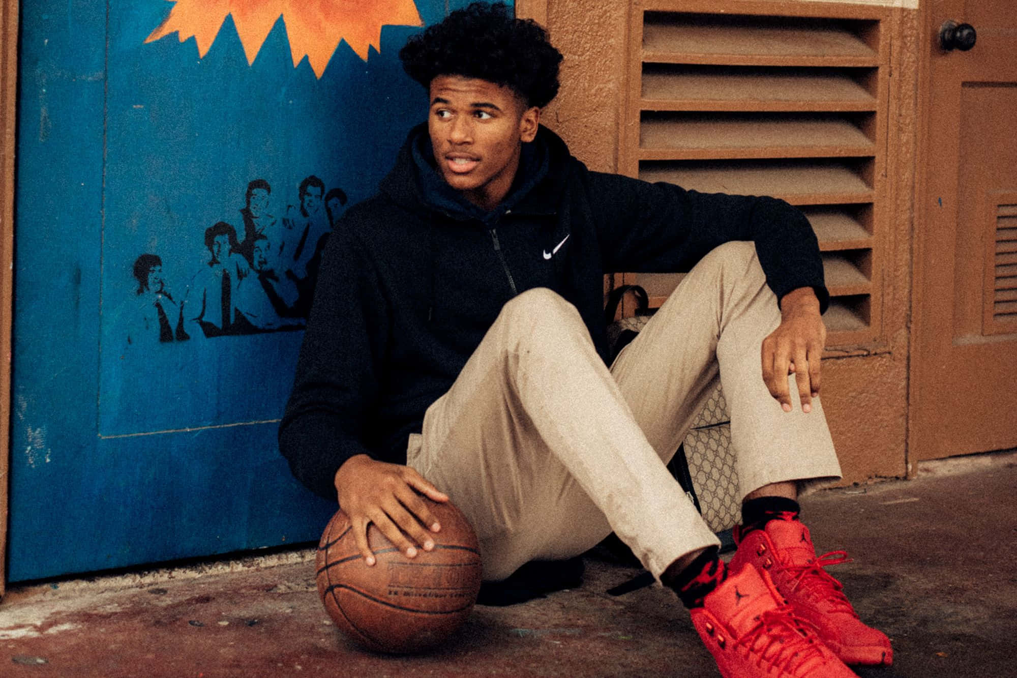 Jalen Green 桌布 壁紙 — 一個穿著紅色運動鞋的年輕籃球運動員，靠著藍色塗鴉牆，隨意地坐著，手持一個舊籃球。