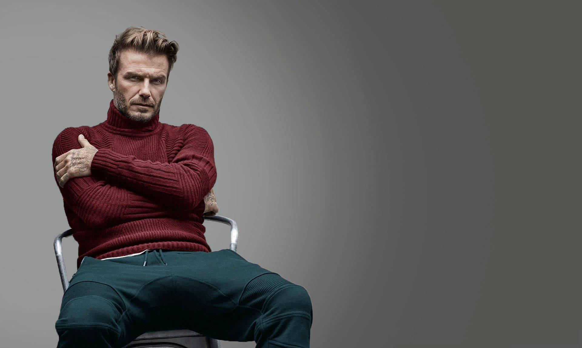 David Beckham 桌布 壁紙 — 一個穿著紅色高領毛衣和綠色褲子的男人自信地坐在椅子上，背景是純灰色。