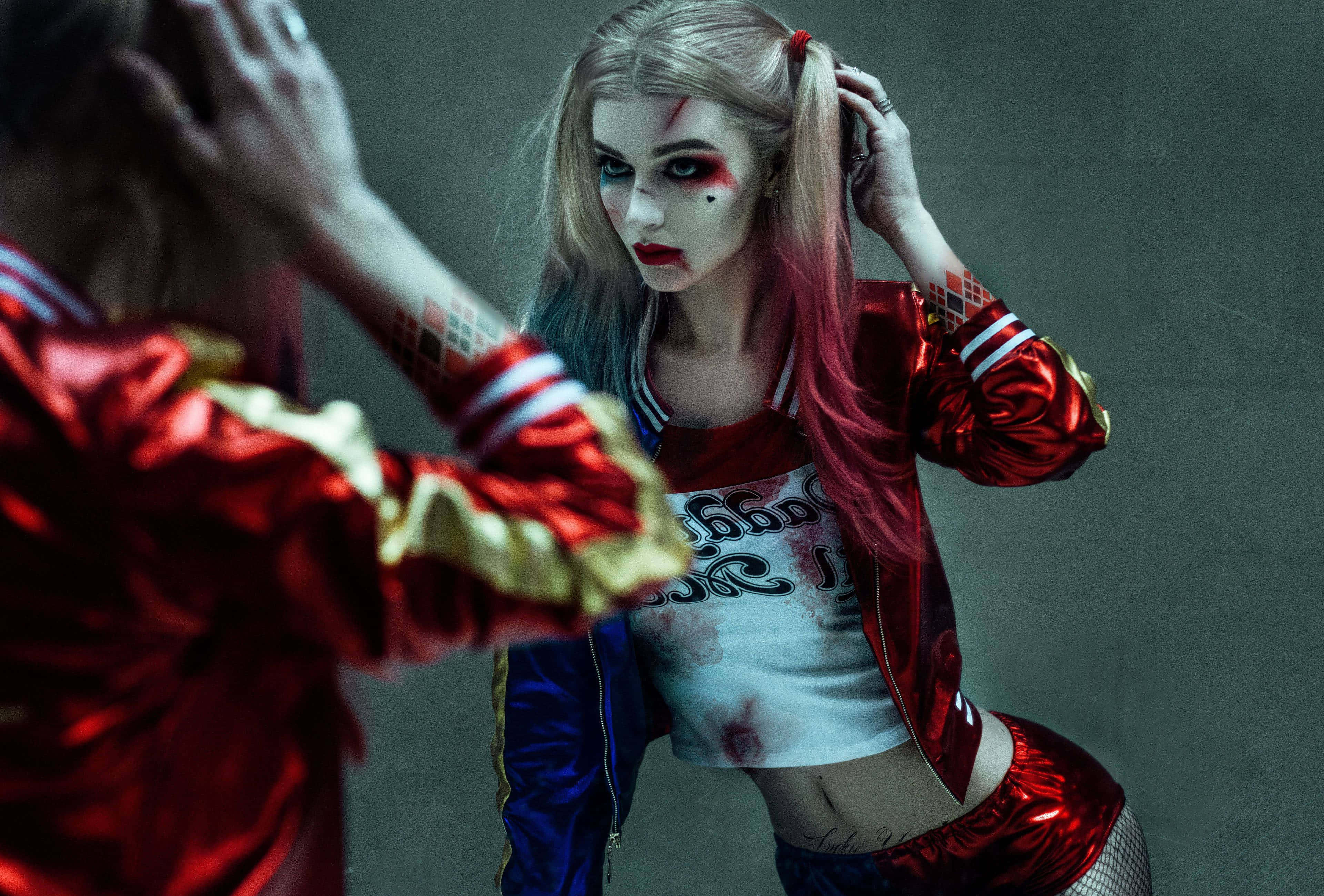 Harley Quinn Cosplay 桌布 壁紙 — 一個穿著紅藍服裝和化妝的哈莉·奎茵角色扮演者，對著灰色牆壁上的鏡子凝視自己的倒影。