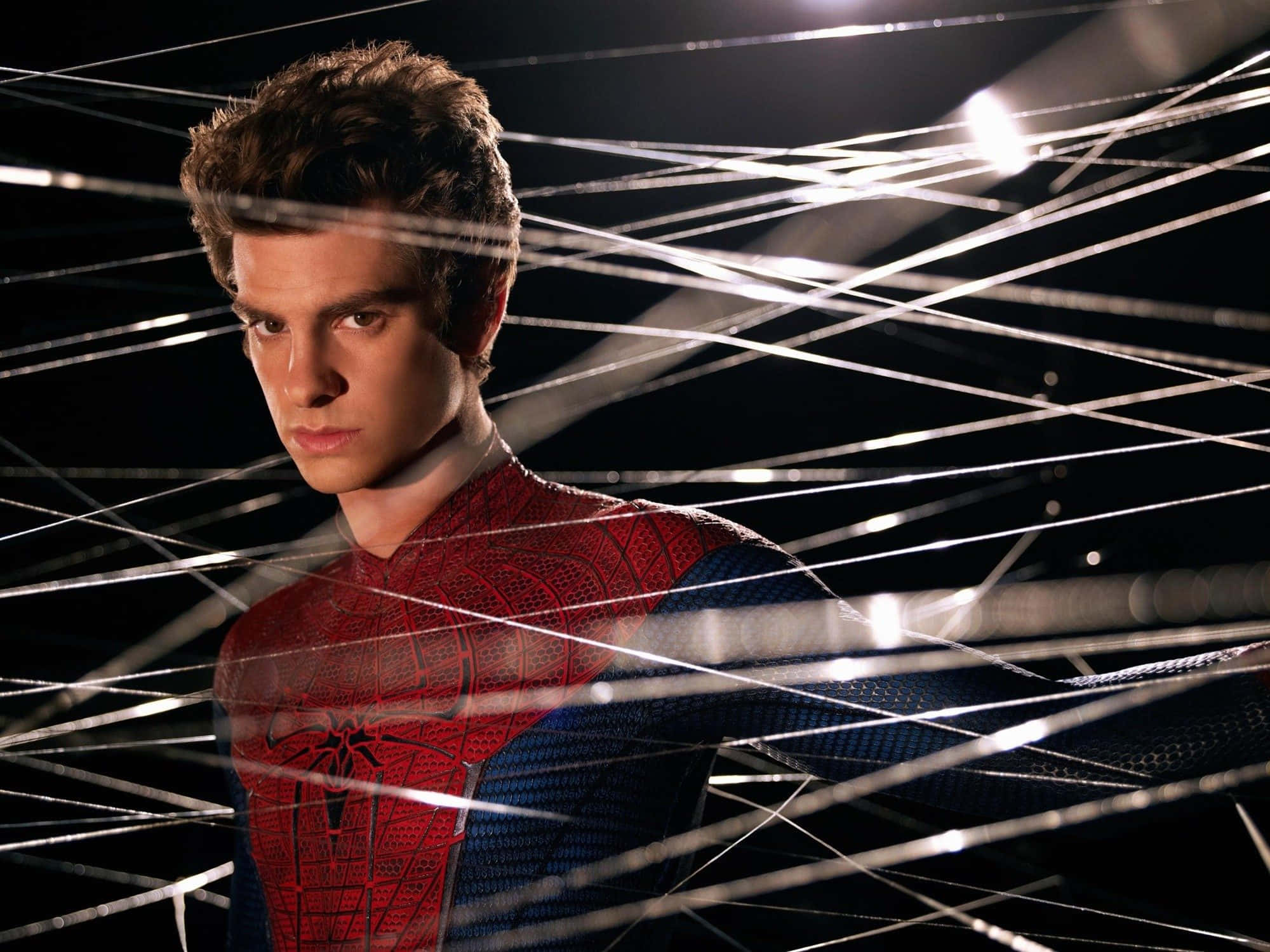 Andrew Garfield Spider Man 桌布 壁紙 — 一個穿著紅藍蜘蛛服的年輕人站在黑暗背景中發光的白色蜘蛛網線之間。