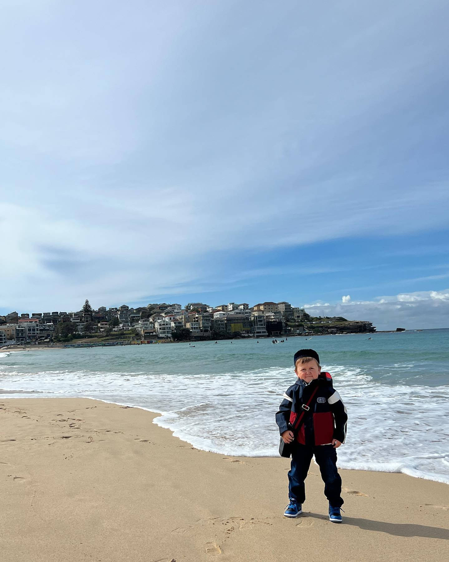 海灘 桌布 壁紙 — 一個穿著紅黑外套的小男孩站在沙灘上，背景有海浪和沿海小鎮，天空部分多雲。