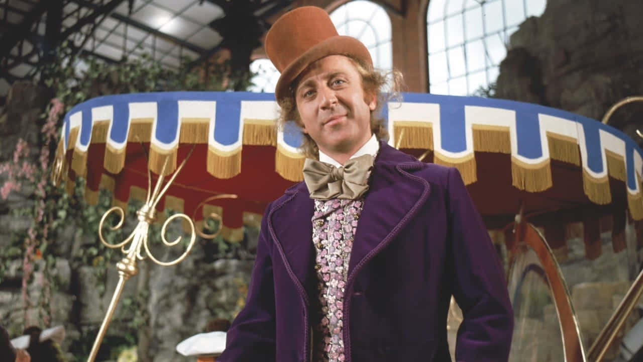 Willy Wonka 桌布 壁紙 — 一個穿著紫色外套和高帽的古怪男人，站在一個夢幻、燈火通明的大廳裡，旁邊是一輛糖果主題的馬車。