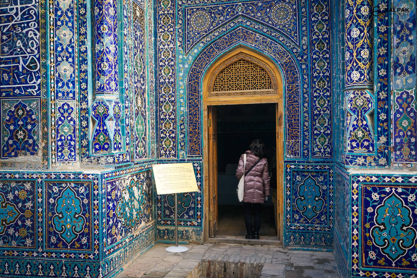 Uzbekistan 桌布 壁紙 — 一個穿著紫色外套的人站在一座充滿活力的藍色瓷磚伊斯蘭建築的門口，該建築帶有複雜的幾何和花卉圖案。