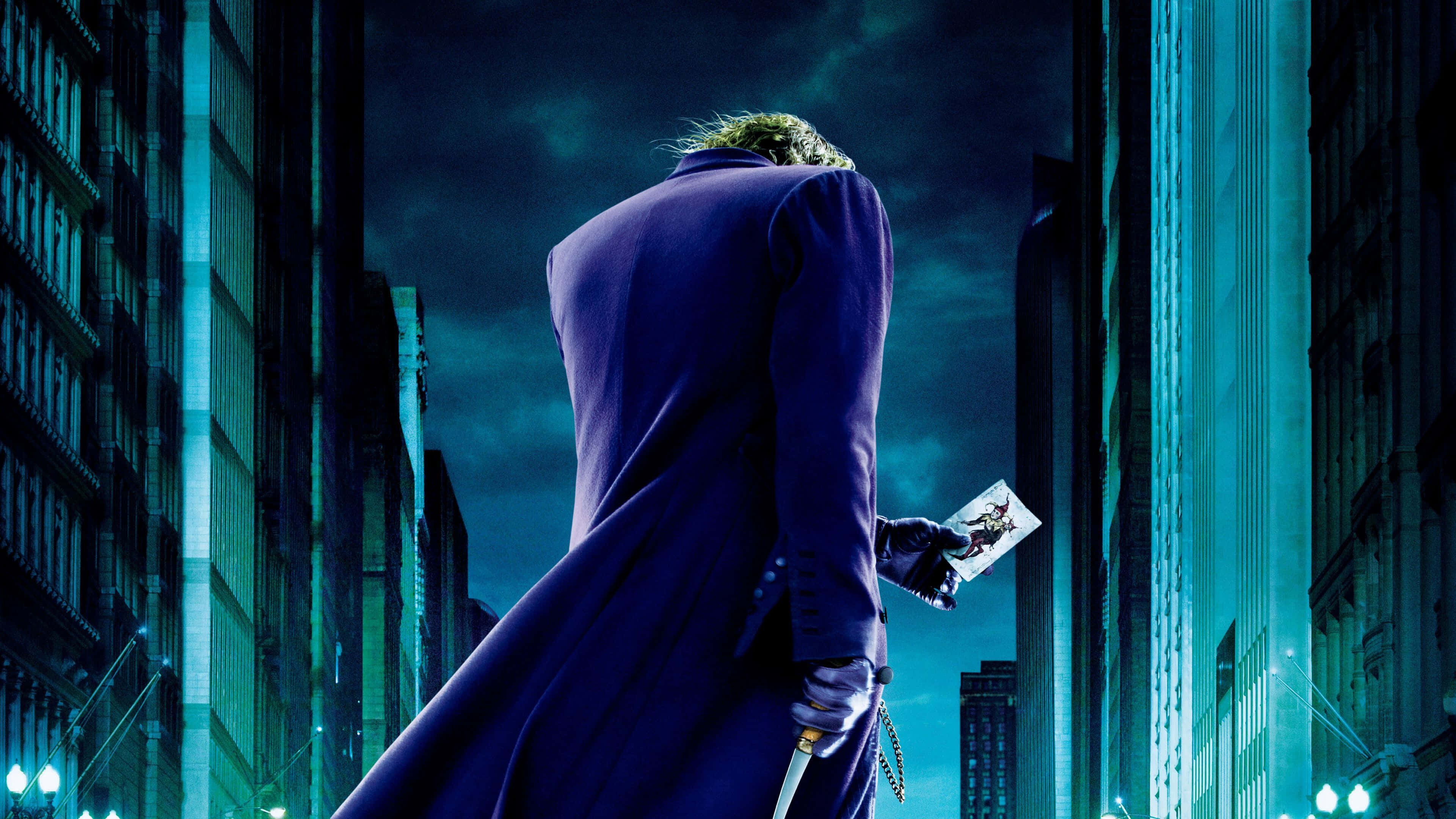 Dark Knight Joker In 4K Ultra HD 桌布 壁紙 — 一個穿著紫色外套的威脅人物站在高聳的城市建築之間，在深藍綠色的天空下拿著一張撲克牌。