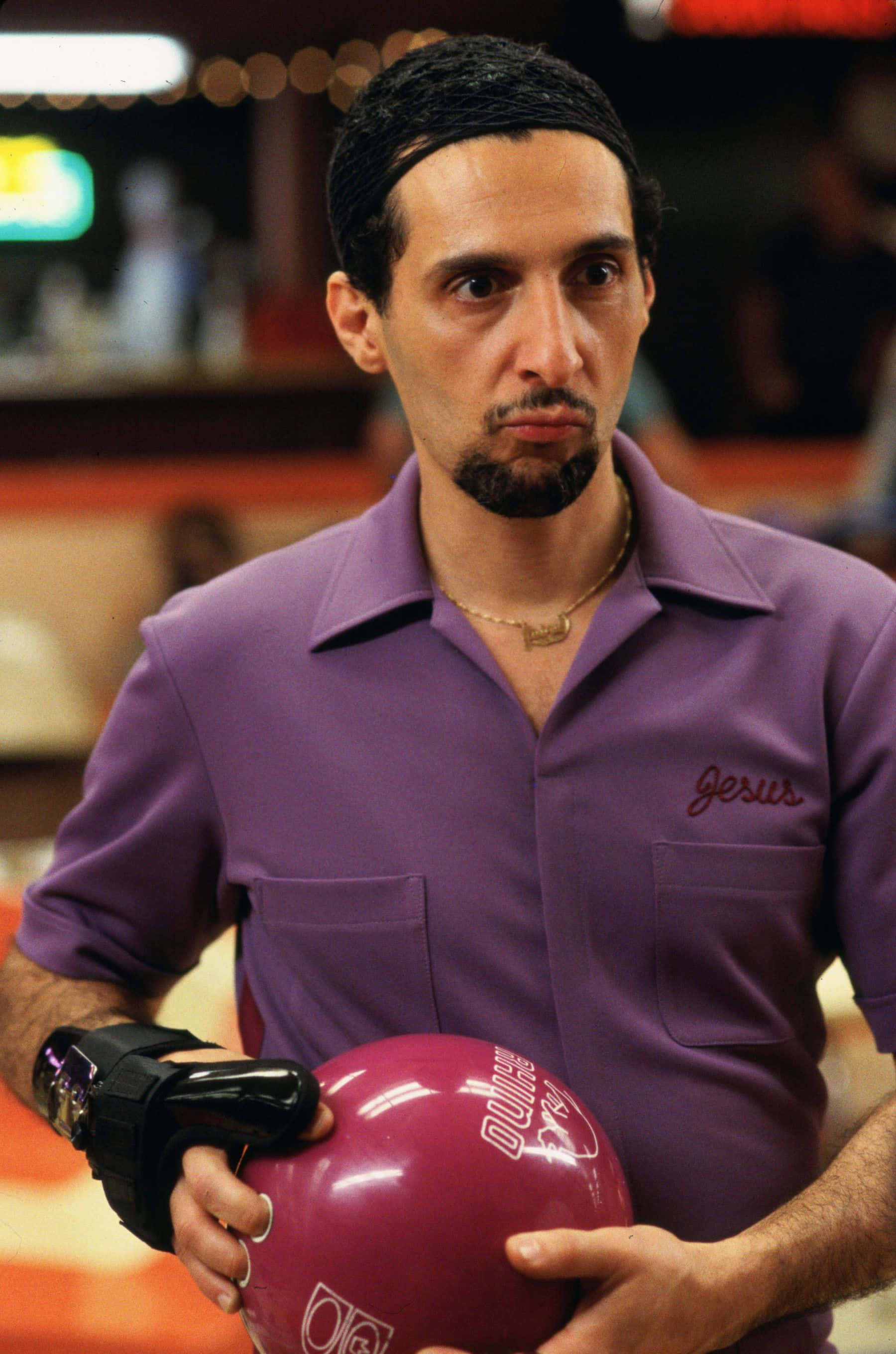 John Turturro 桌布 壁紙 — 一個穿著紫色襯衫的男人在光線昏暗的保齡球館裡，表情嚴肅地拿著一個粉紅色的保齡球。