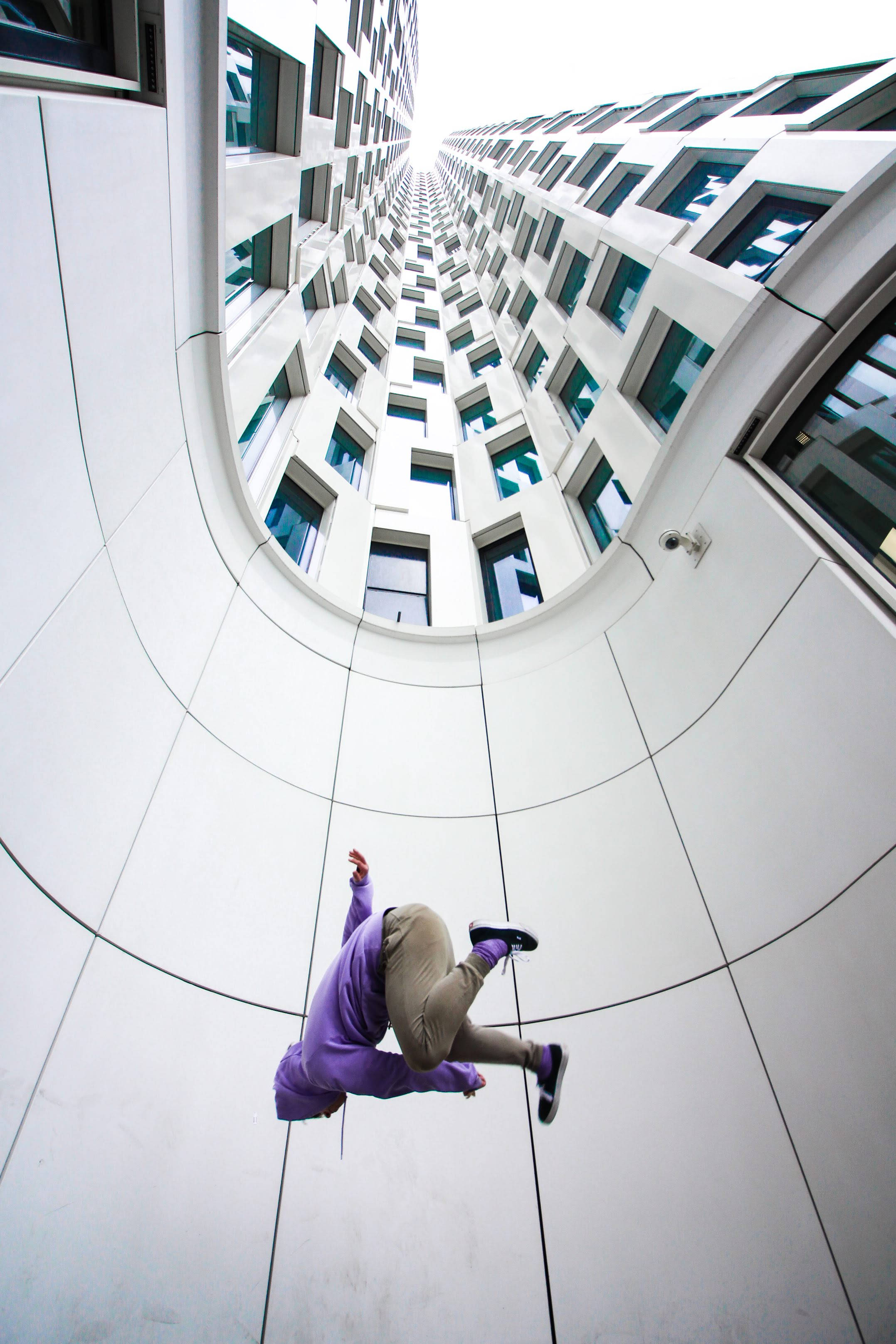 Parkour 桌布 壁紙 — 一個穿著紫色連帽衫的人在高大的白色建築物之間騰空跳躍，建築物有青綠色窗戶，天空明亮。
