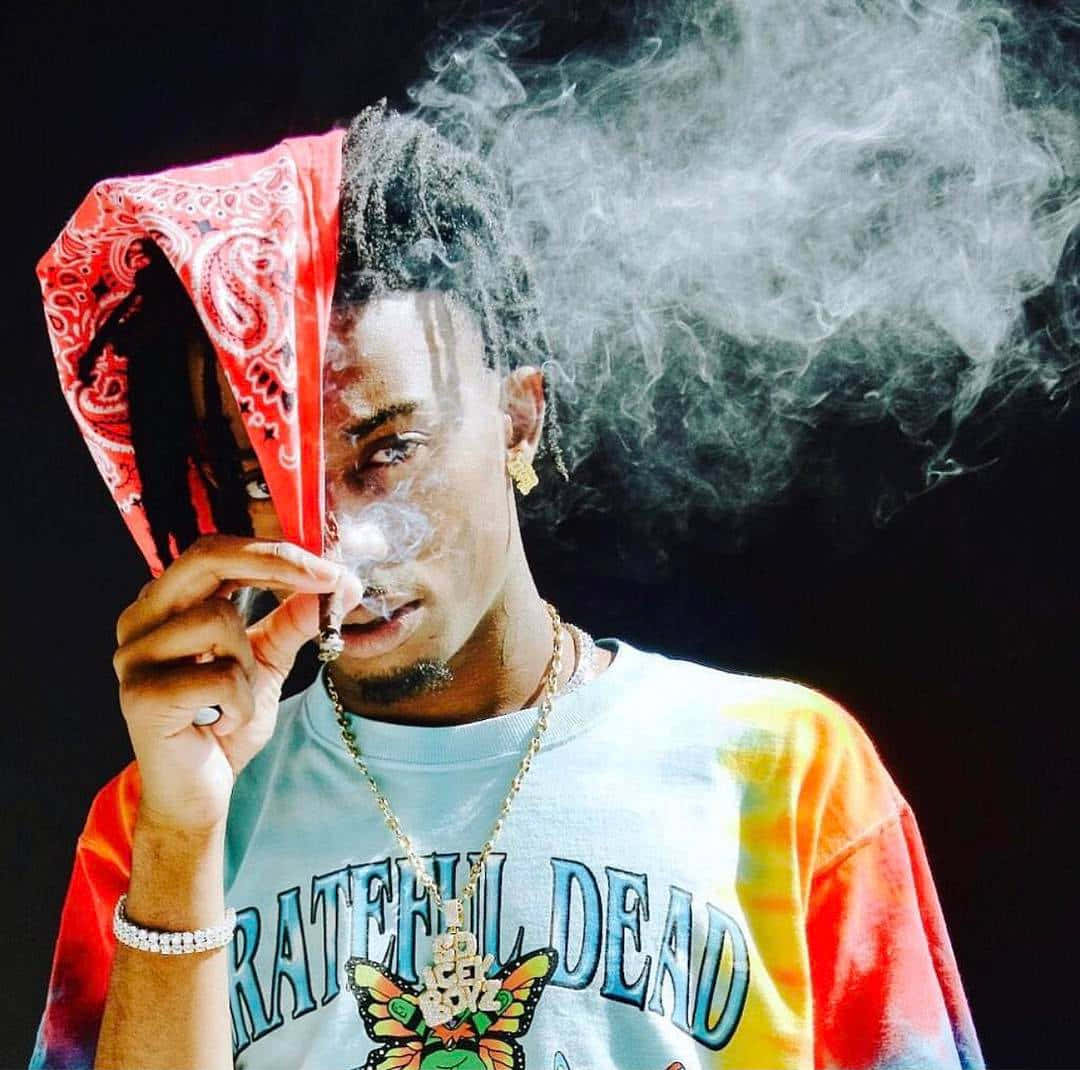 Playboi Carti HD 桌布 壁紙 — 一個穿著紮染Grateful Dead T恤的年輕男子呼出煙霧，半邊臉被紅色頭巾遮蓋。