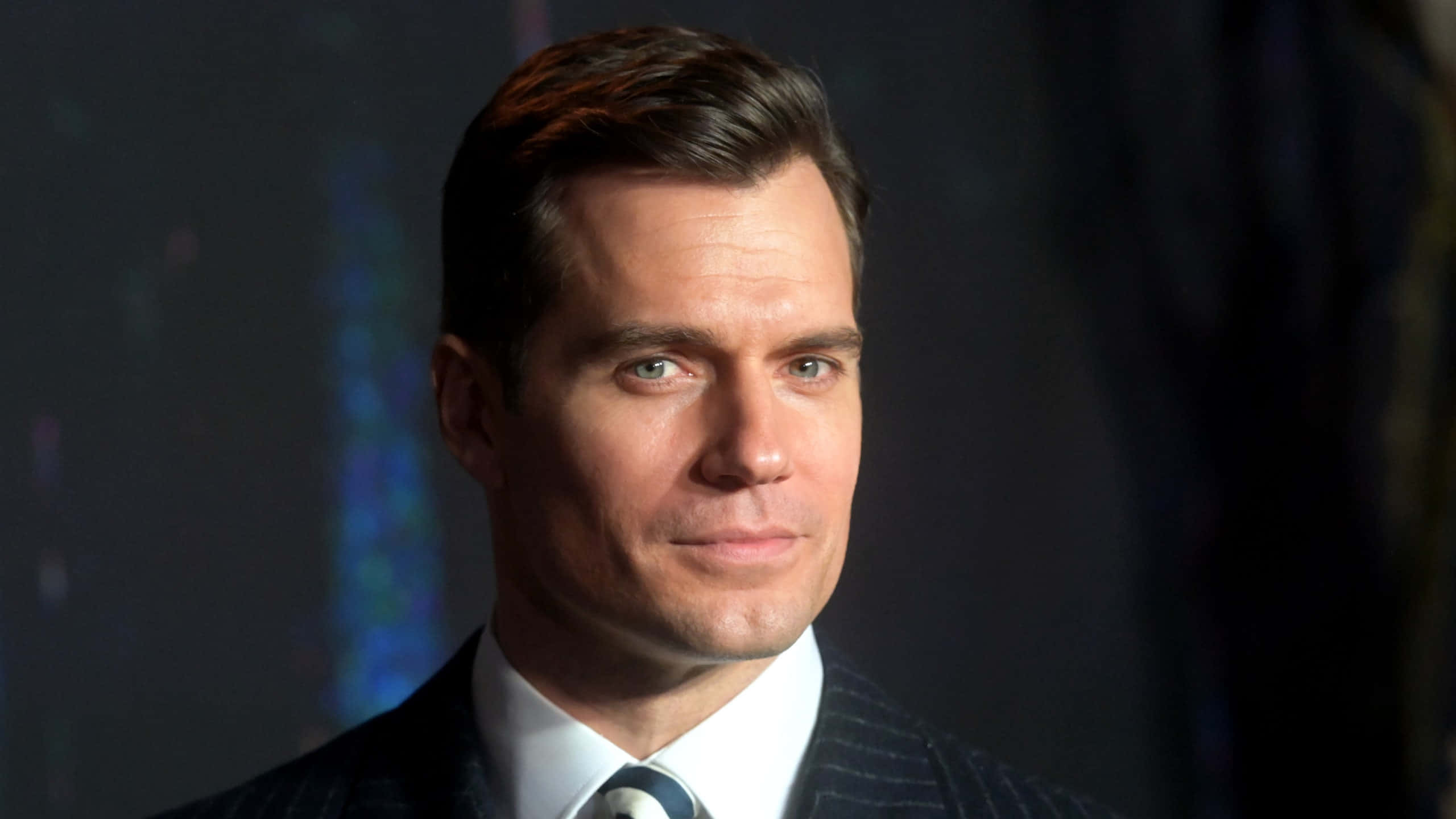 Henry Cavill 桌布 壁紙 — 一個穿著細條紋西裝的男人在黑暗模糊背景下的特寫肖像，帶著微妙微笑。